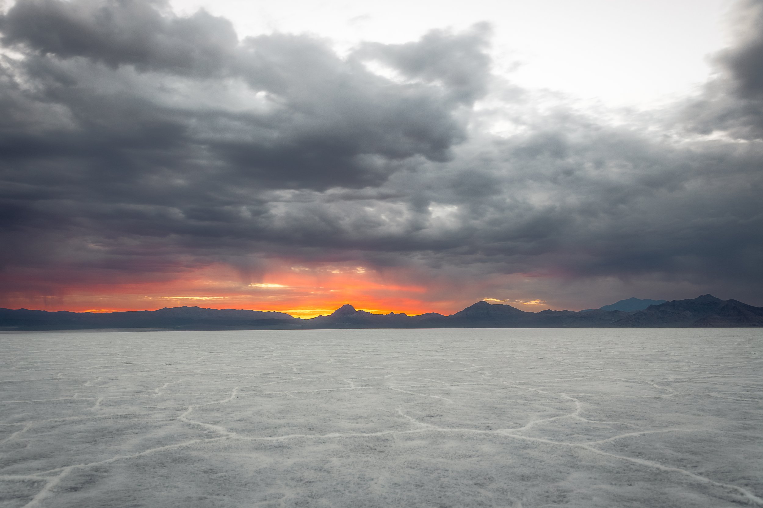 A landscape photograph of a sunset over a mountain range with a body of water in the foreground. The sky is filled with dark, heavy clouds, with the horizon showing hues of orange, pink, and yellow.