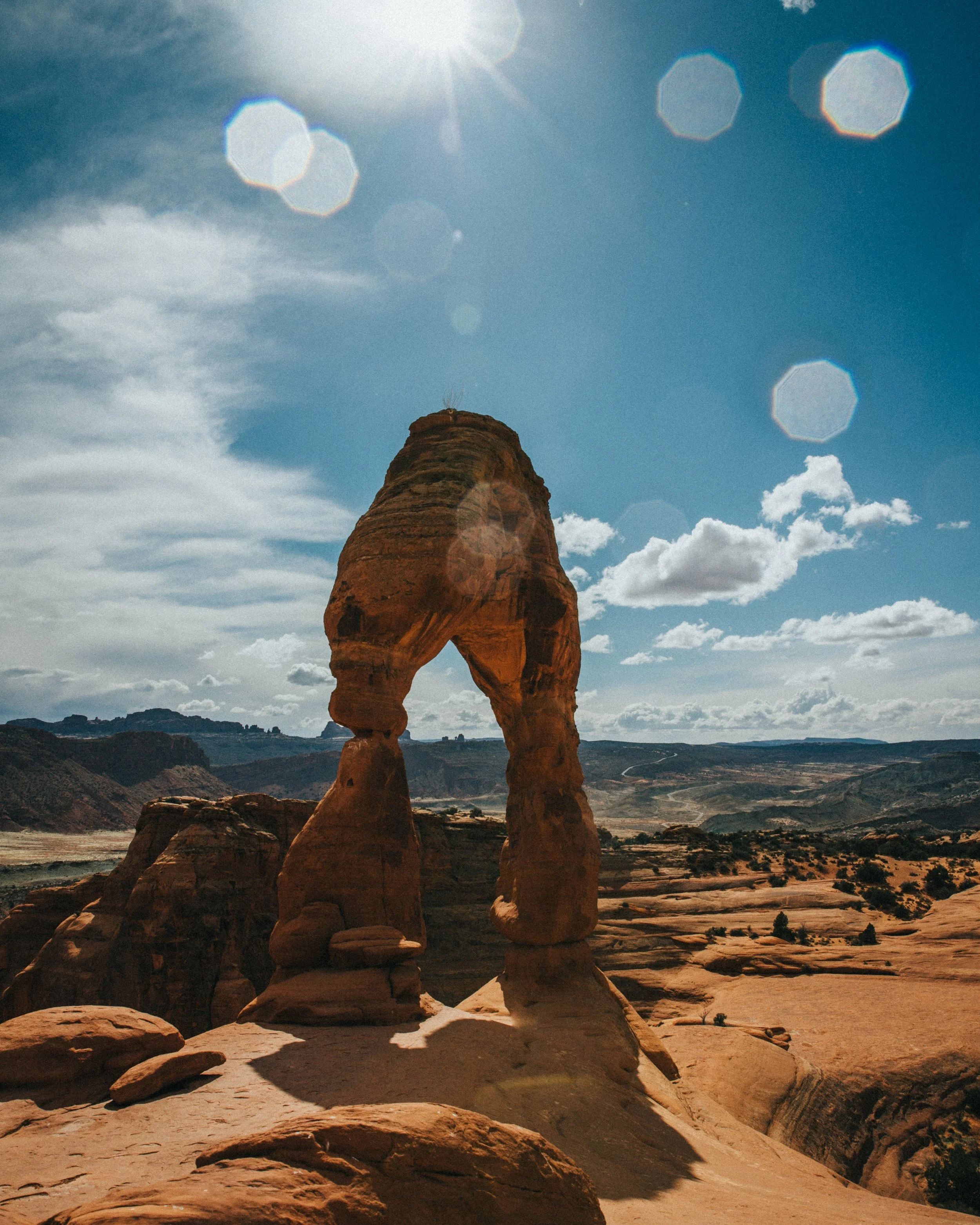 A rock formation known as Delicate Arch located in Arches National Park, Utah, under a sunny sky with some clouds and lens flare effects.