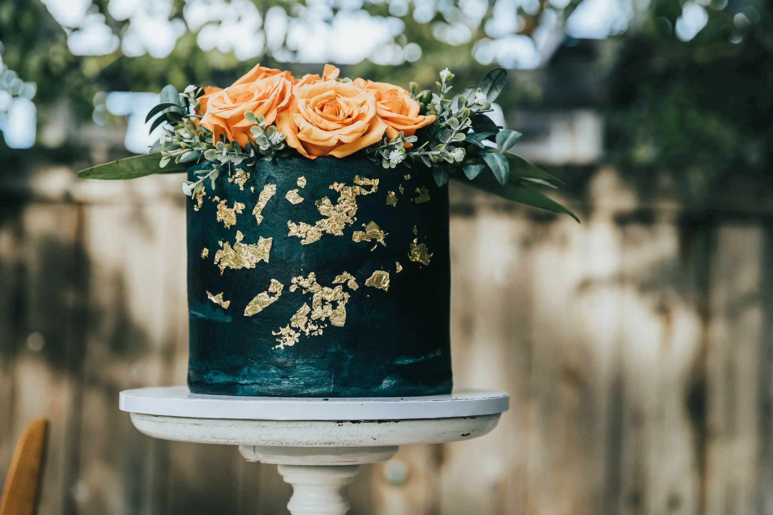 A flower arrangement with orange roses and greenery in a dark green vase with gold leaf patterns, placed on a white pedestal outdoors.