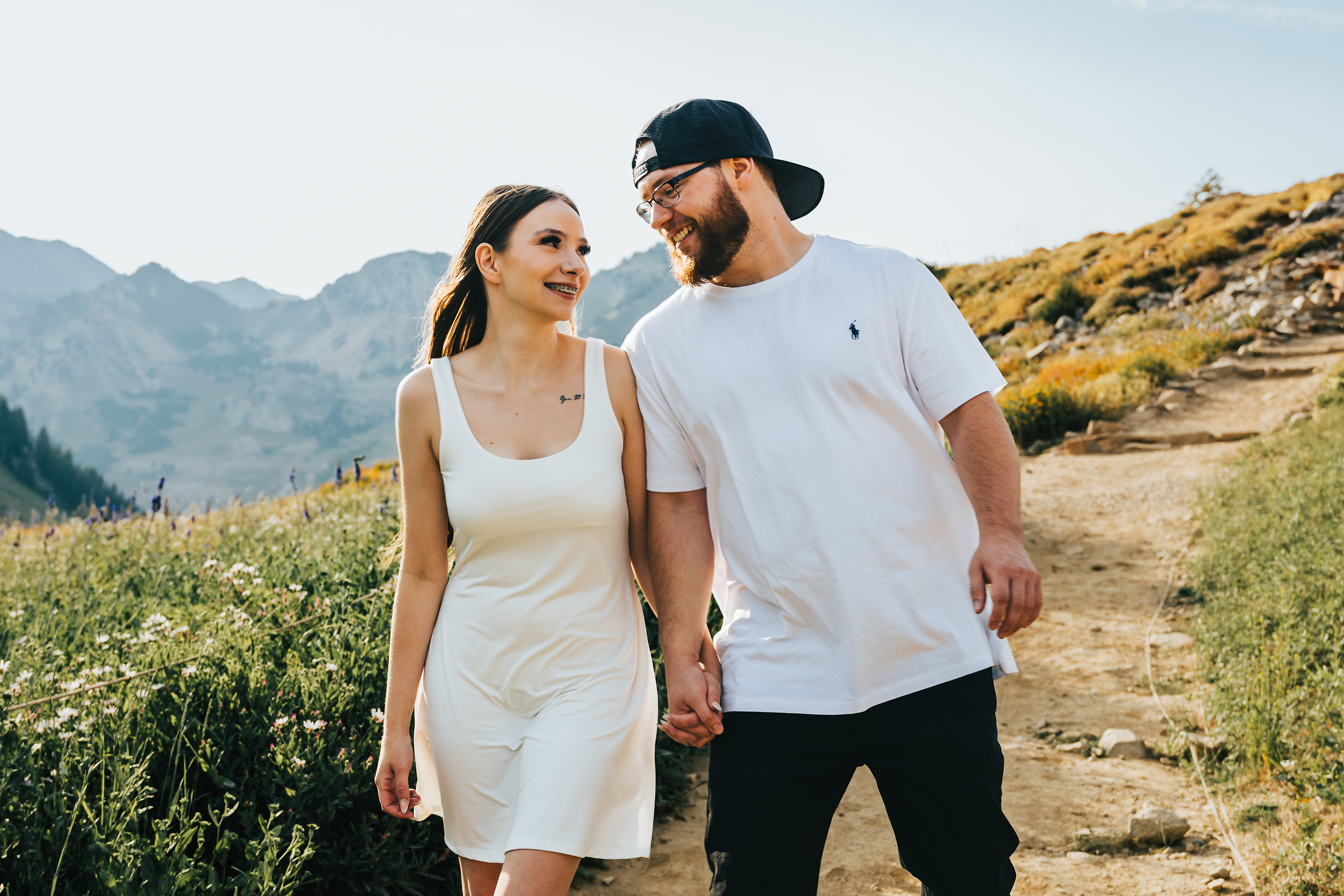 A young woman and man holding hands and walking outdoors along a mountain trail with wildflowers and hills in the background, smiling at each other.
