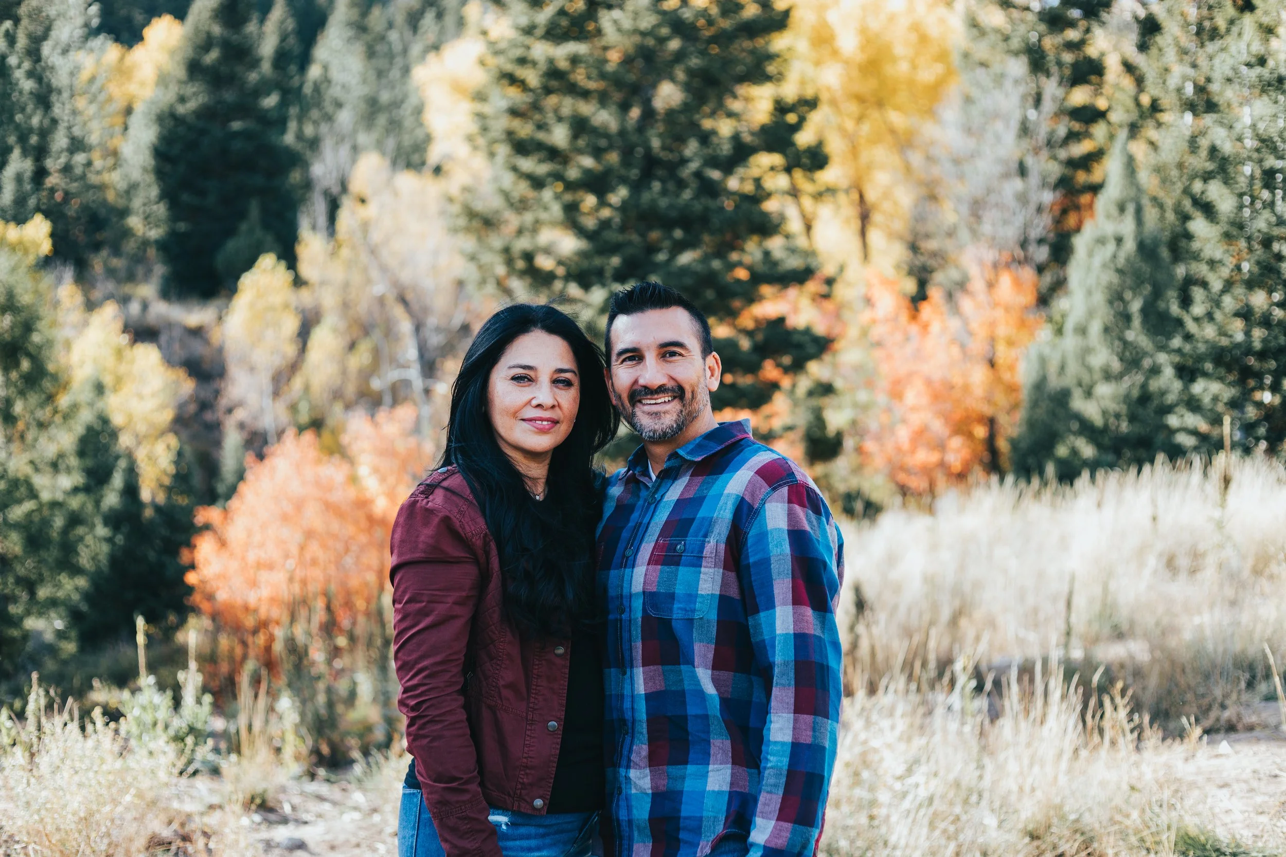 A smiling couple standing outdoors in a fall forest with colorful autumn trees in the background.