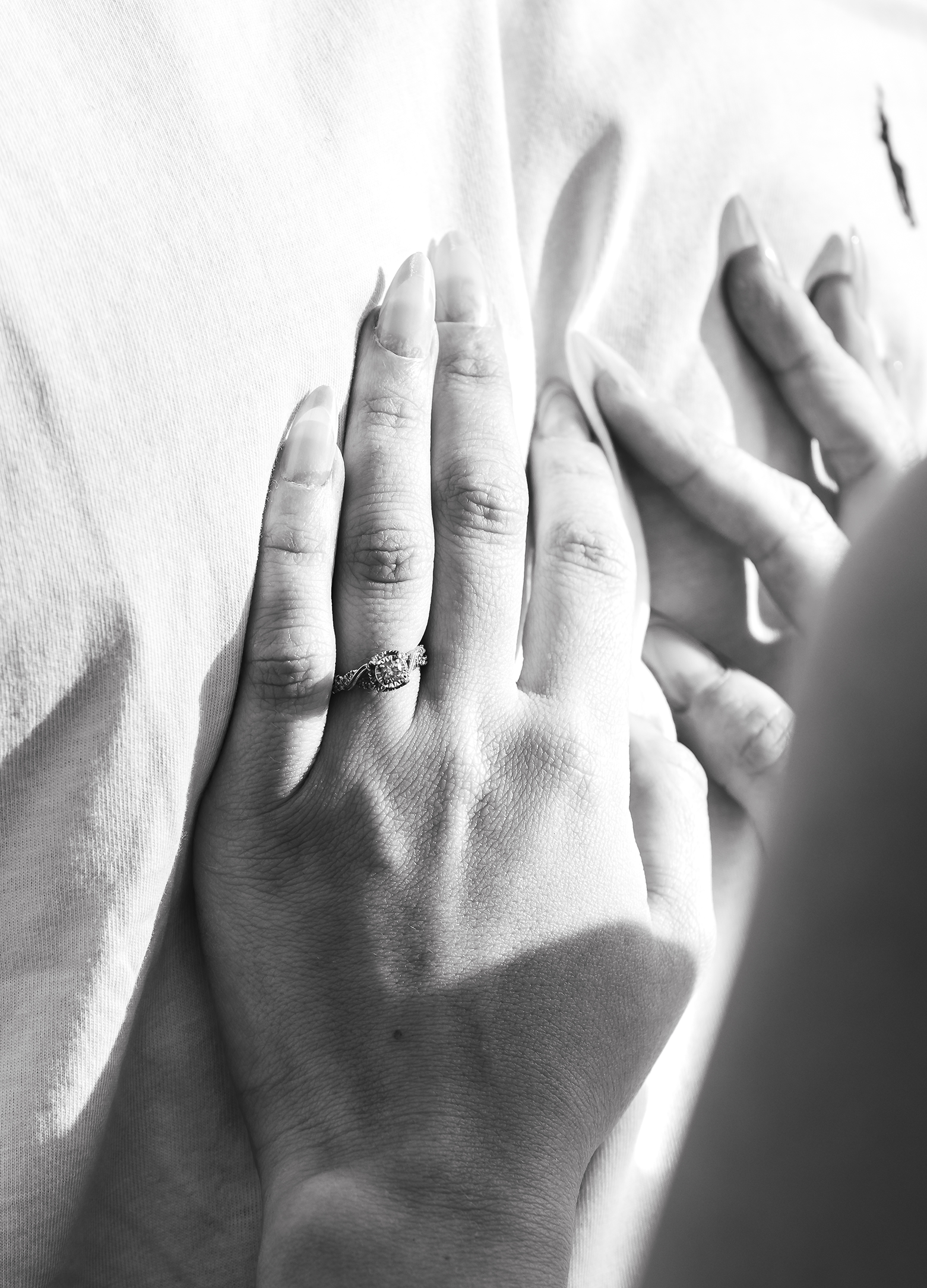 Close-up black-and-white photo of a person's hand gently resting on skin, wearing an engagement ring with a large diamond, on a smooth fabric background.