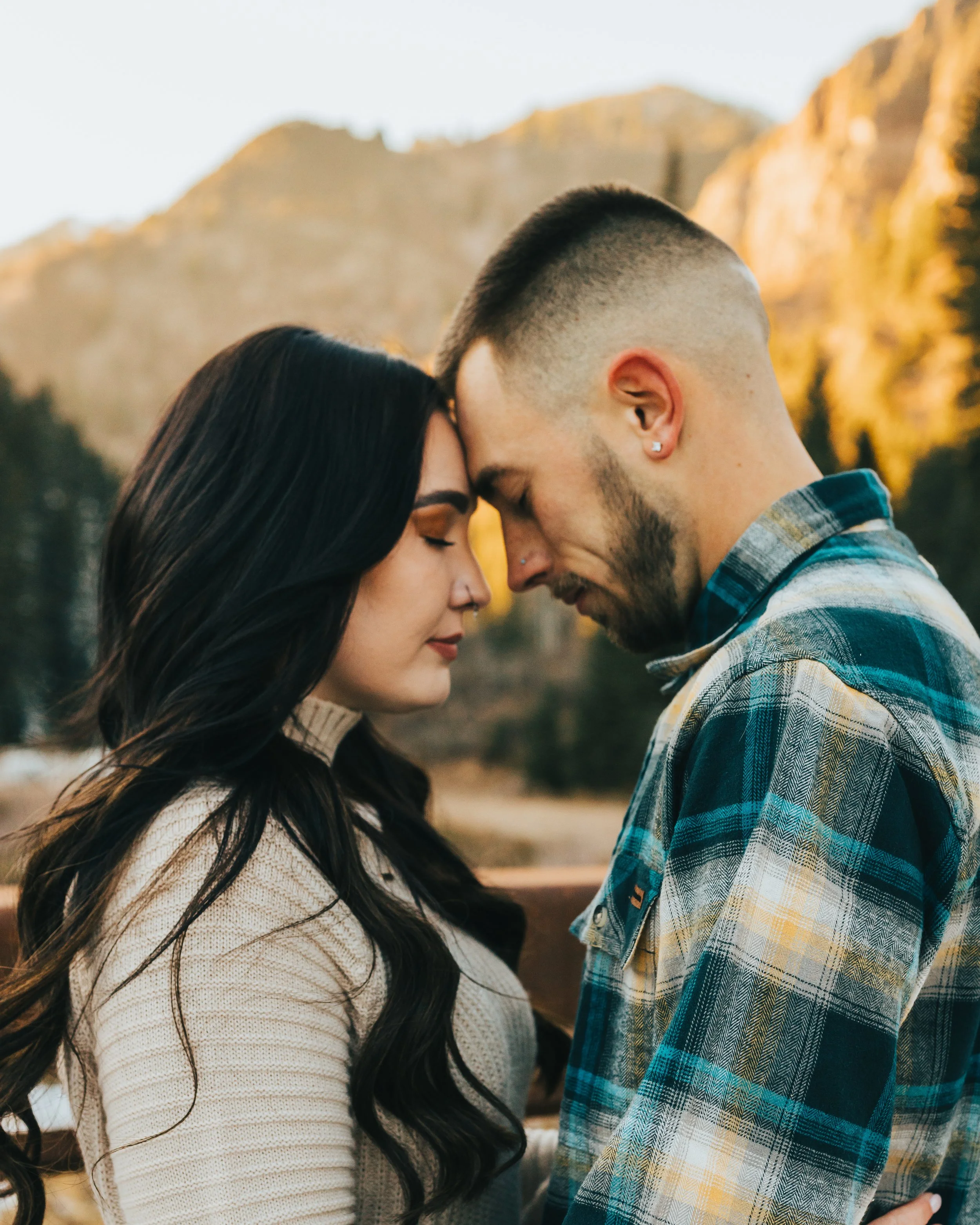 A young couple with foreheads touching and eyes closed in an outdoor setting during sunset with mountains in the background.