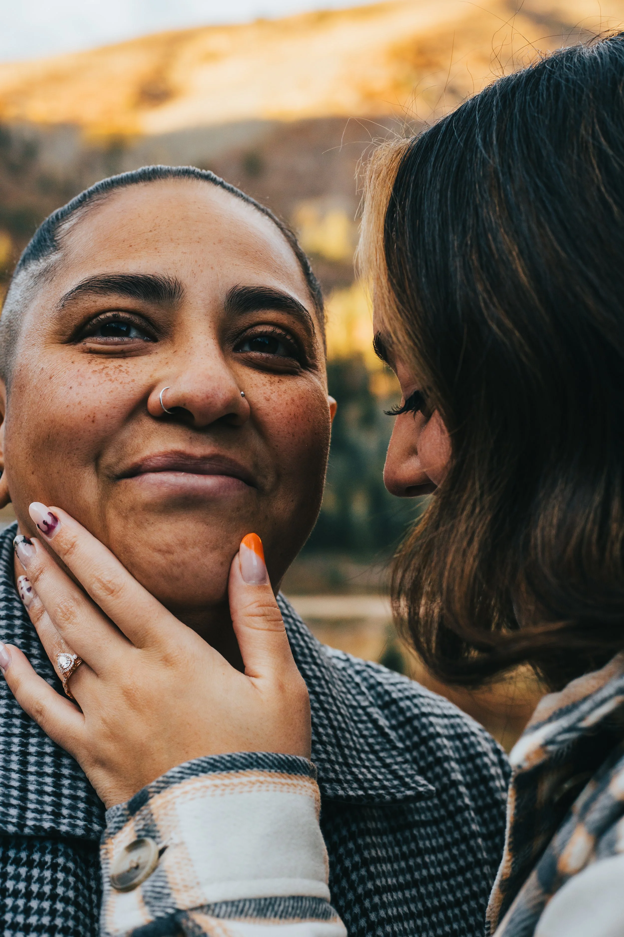 Close-up of two women, one with short dark hair and freckles, and the other with long brown hair, sharing an intimate moment outdoors during sunset.