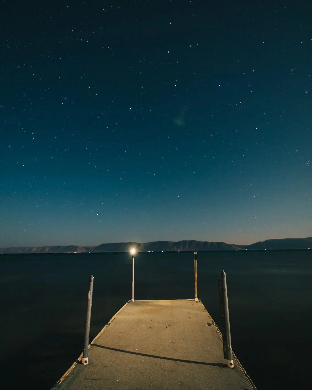 Nighttime view of a dock extending into a dark lake, with hills in the background and a star-filled sky overhead.