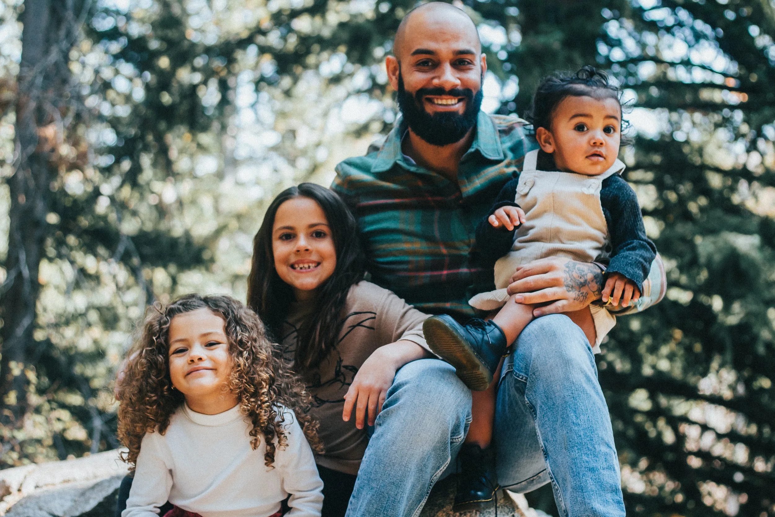 A group of children and an adult man outdoors in a forested area, smiling and sitting on rocks, with tall trees in the background.