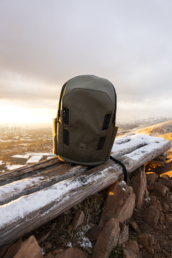 Gray backpack resting on a snow-dusted wooden bench on a hilltop at sunset, overlooking a cityscape.