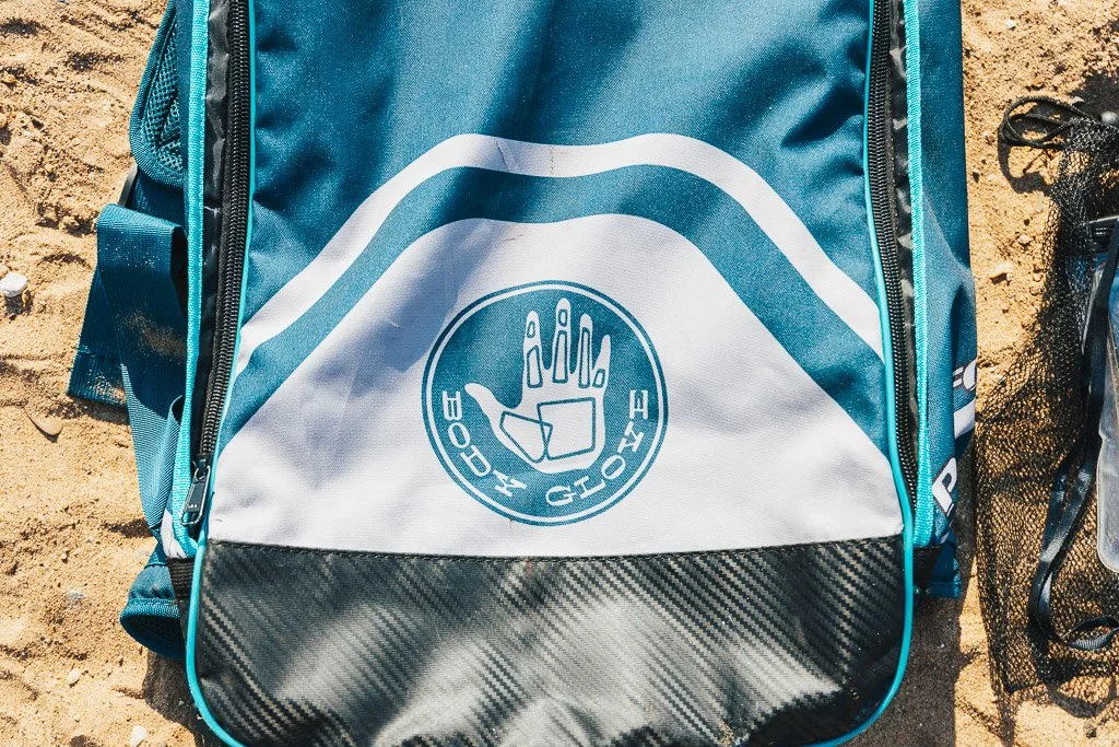 A blue and gray backpack with a black carbon fiber-patterned lower section, placed on sandy ground. The backpack features a white circular logo with a stylized hand making a stop gesture, encircled by the words "HOPE" and "GLOBE," and has zippers on the sides.