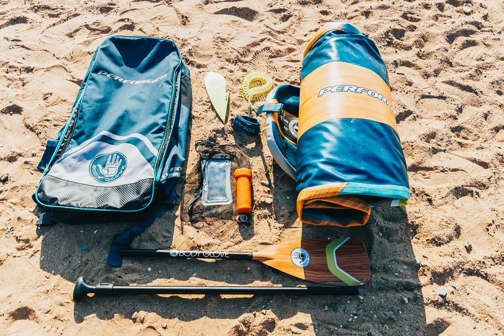 Beach gear including a blue backpack, an ice cream cone, a blue and yellow rolled-up beach mat, a small orange and black flashlight, a blue silicone phone holder, a white and green cup, a paddleboard paddle, and a blue and yellow life vest, arranged on sandy ground.