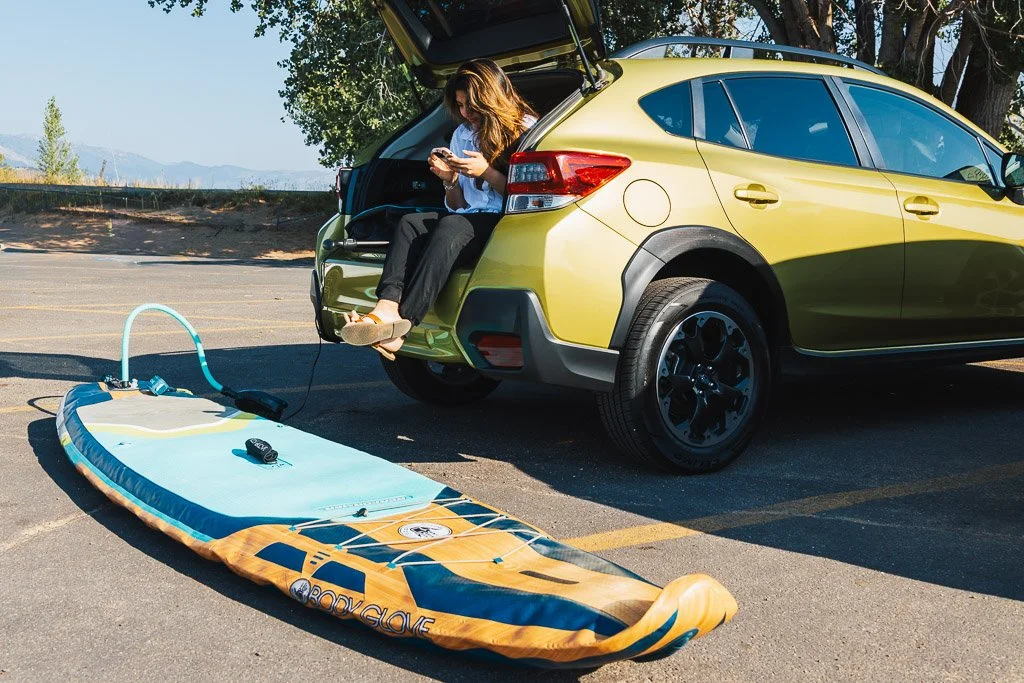 A woman sitting in the open trunk of a yellow hatchback car, looking at her phone, with a paddleboard on the parking lot ground nearby, connected to the car for inflation.
