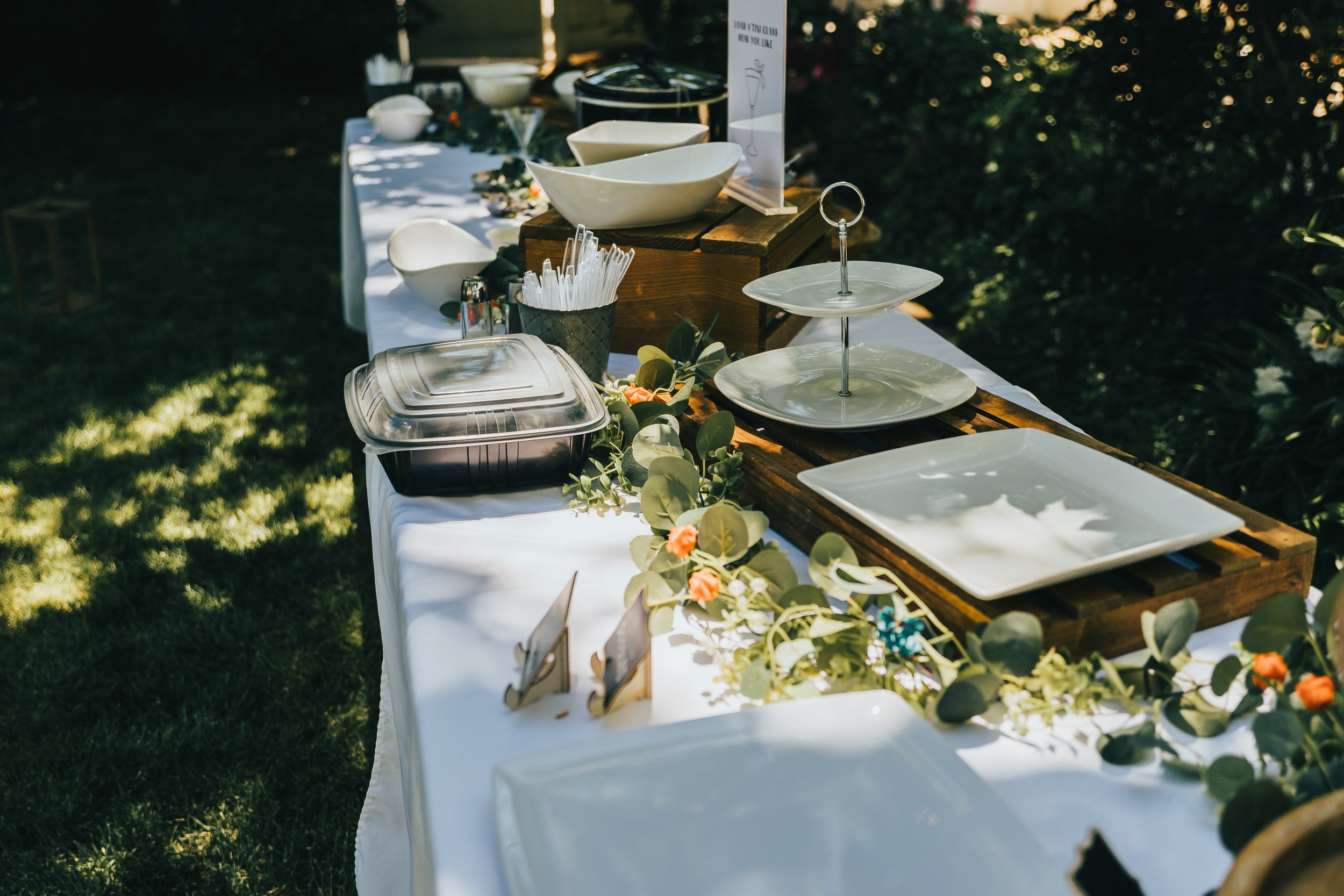 Outdoor buffet table with white dishes, glassware, and greenery, set up in a garden with sunlight filtering through trees.