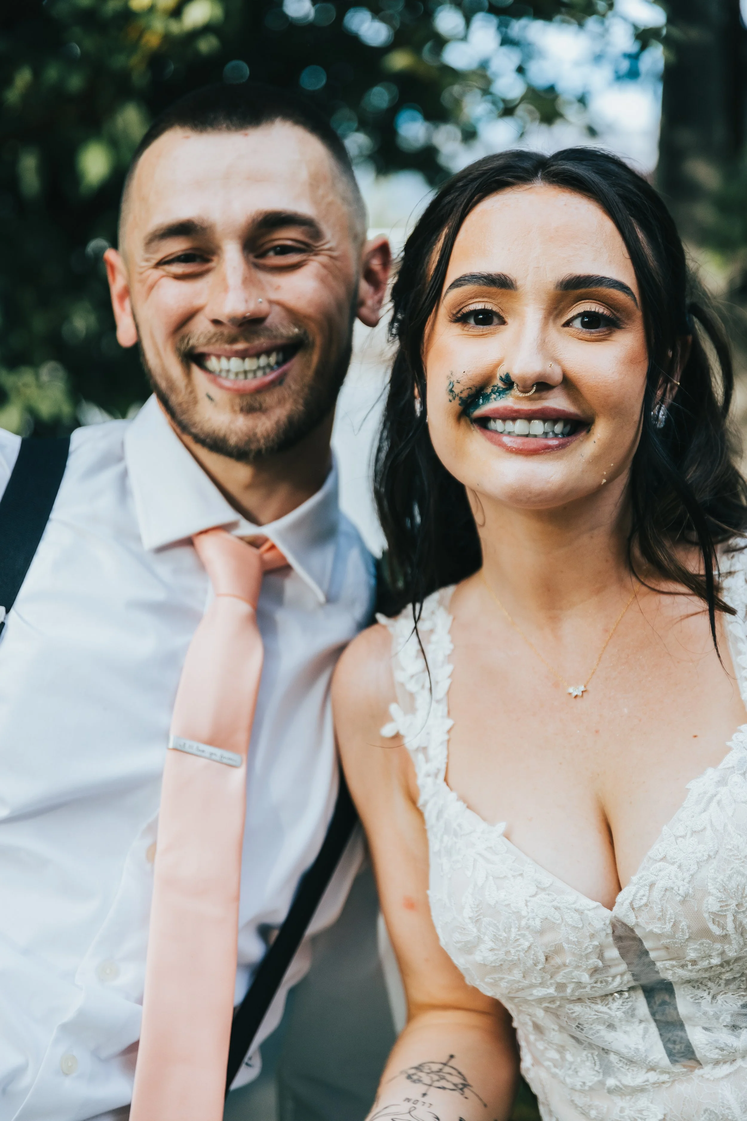 Close-up of a smiling couple at a wedding, the man in a white shirt with suspenders and the woman in a lace white wedding dress, with a tattoo on her arm and blue makeup around her nose.