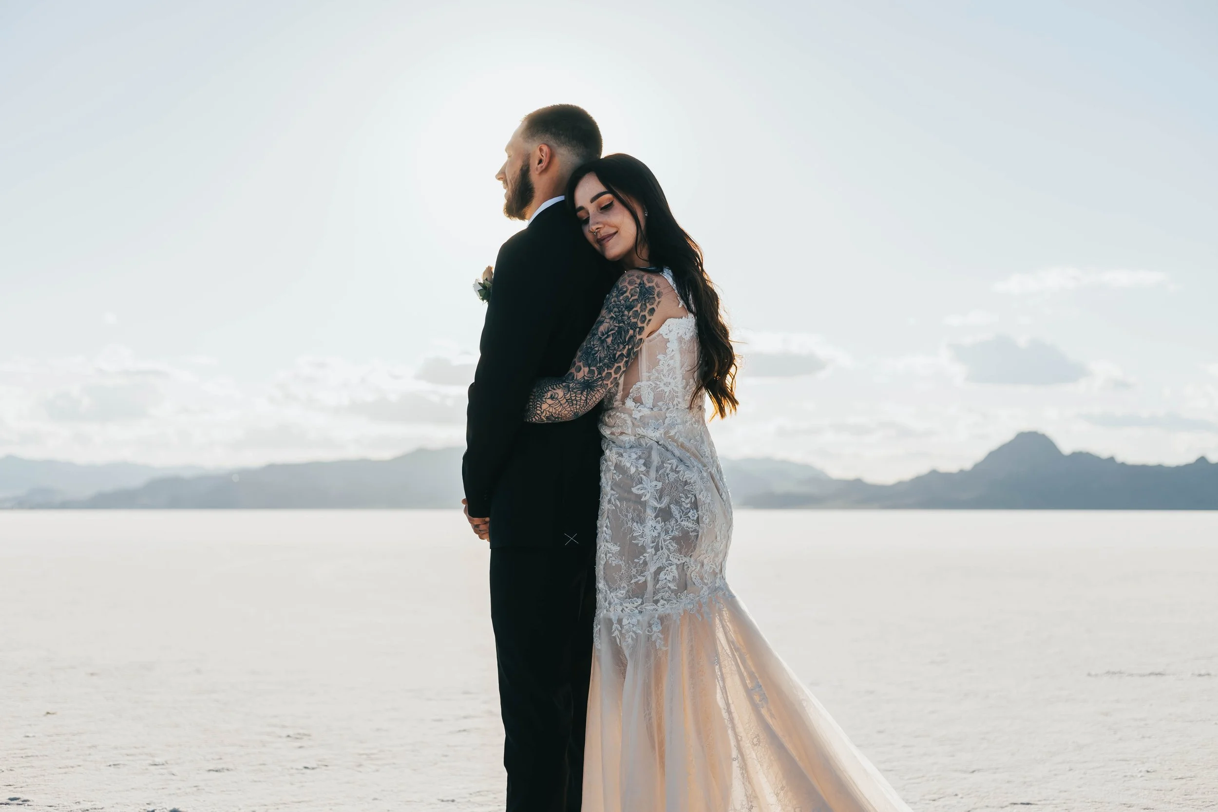 A couple dressed in wedding attire stands closely together on a beach, facing away from the camera, with the bride resting her head on the groom's shoulder during sunset or sunrise.