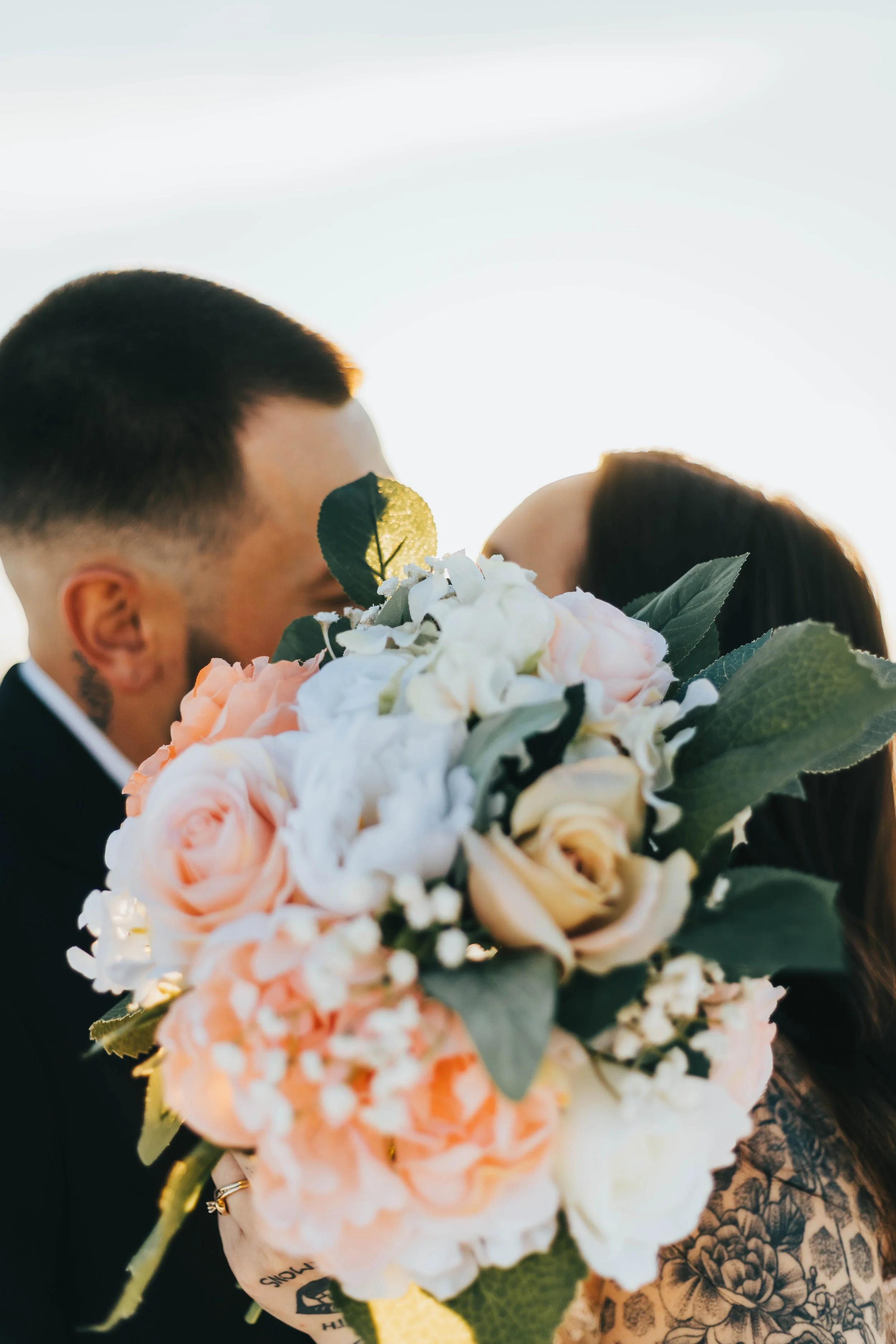 A man and a woman are partially visible, kissing behind a large bouquet of pink and white flowers. The man has short dark hair and tattoos on his neck, and the woman has long dark hair. The scene appears to be outdoors with a bright sky in the backgr