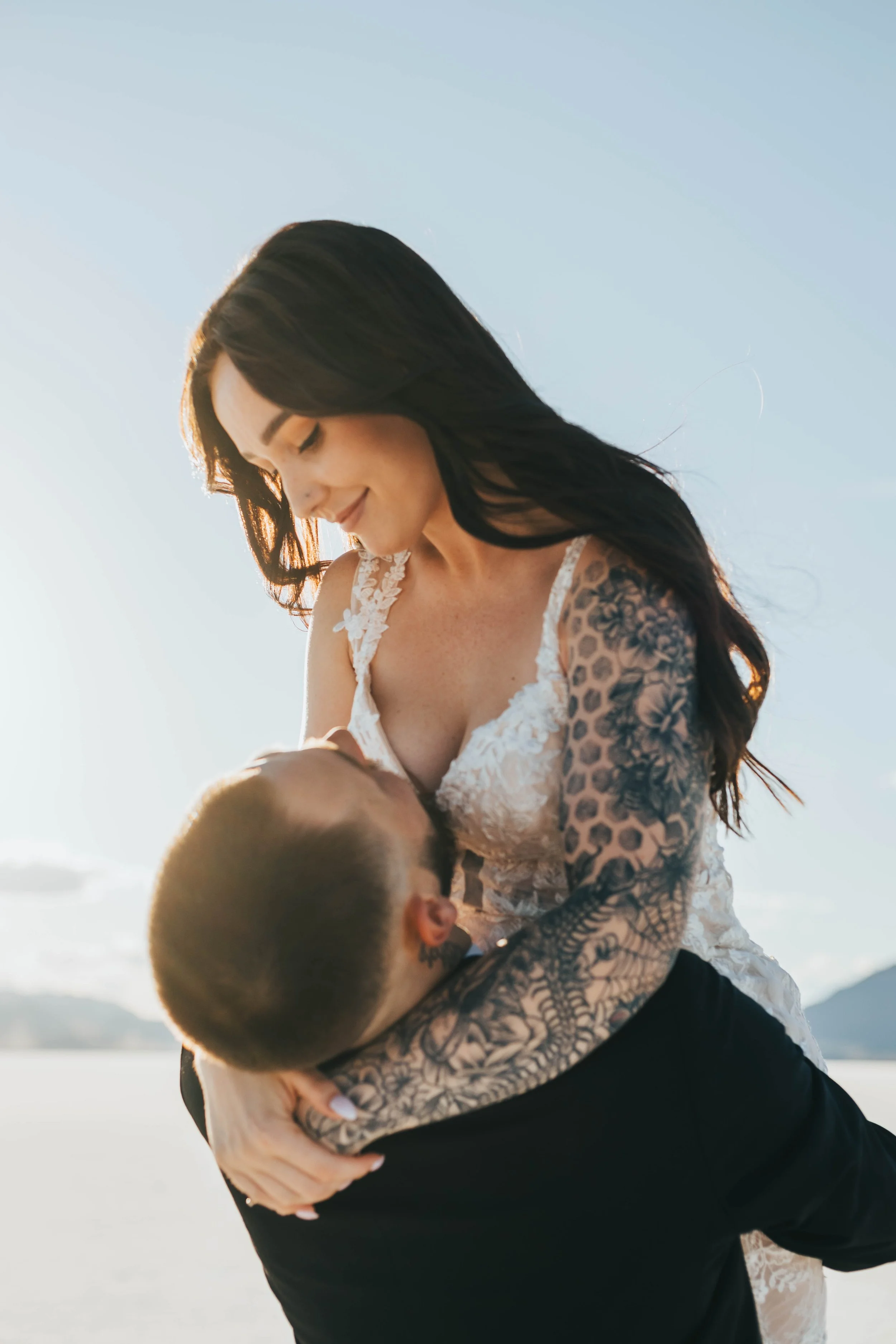 A woman with long dark hair and tattoos on her arms is dancing with a man outdoors during sunset, with water and distant mountains in the background.
