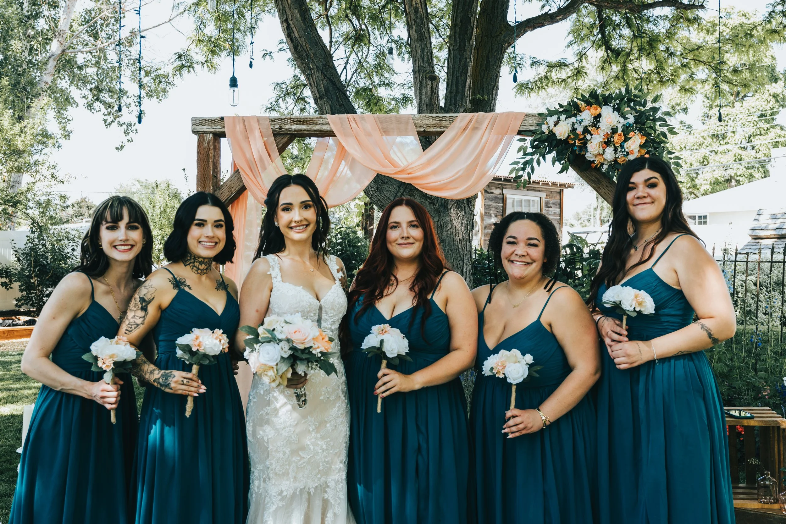 Group of six women in blue dresses and a bride in a white wedding gown posing outdoors under a wooden arch decorated with peach fabric and a floral arrangement, with trees and a house in the background.