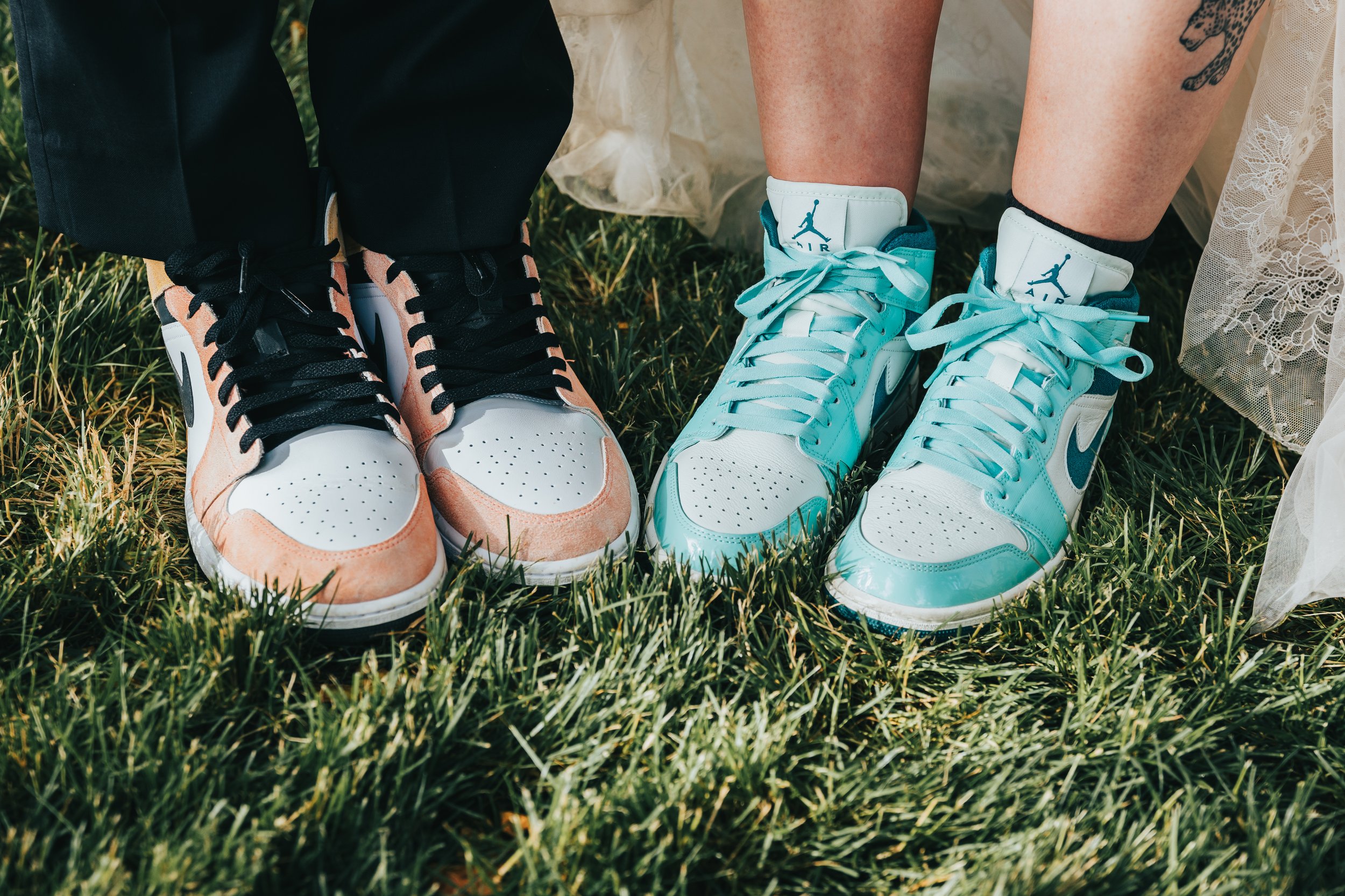 Close-up of two pairs of athletic shoes, one pair pink and black, and the other light blue and white, on grass with legs and a lace-embellished dress visible.