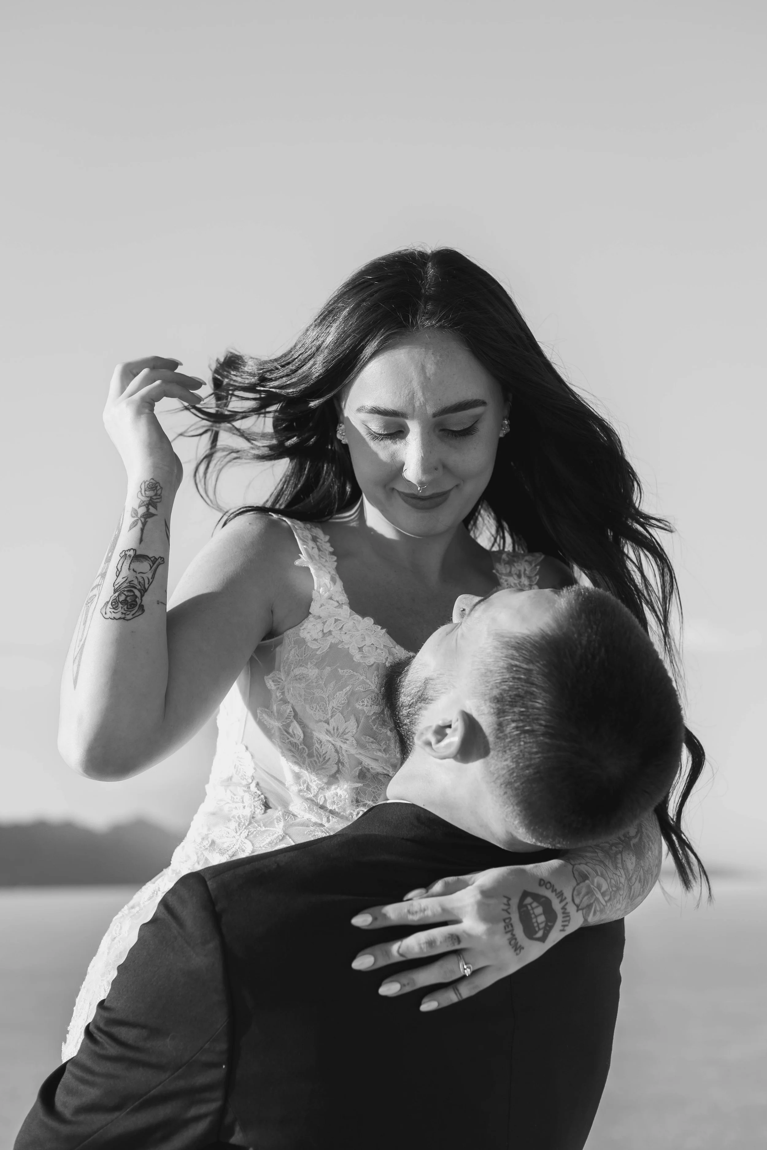 A woman in a wedding dress being lifted by a man, holding him with both hands, outdoors against a clear sky.