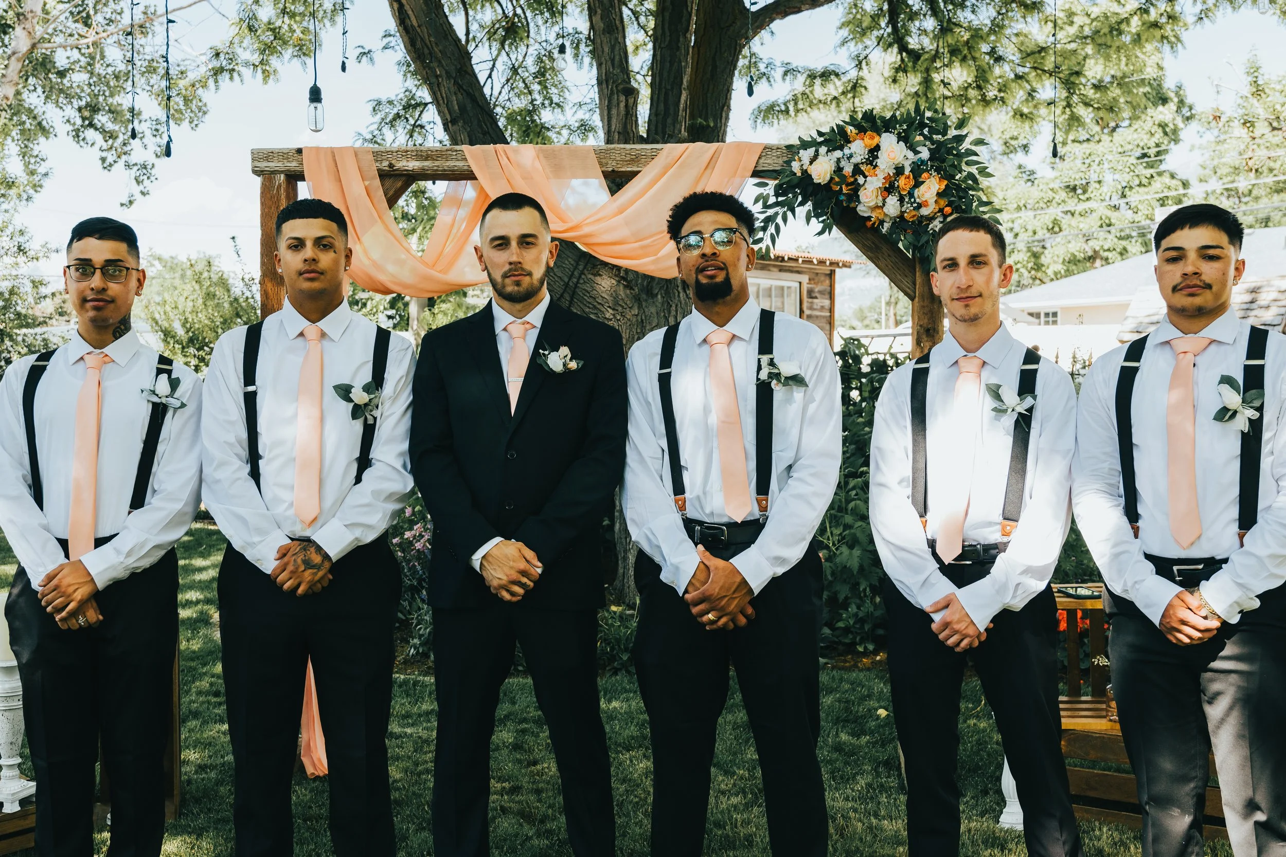 Group of six men dressed in formal attire standing outdoors in front of a decorated wooden arch, with trees and a house in the background.