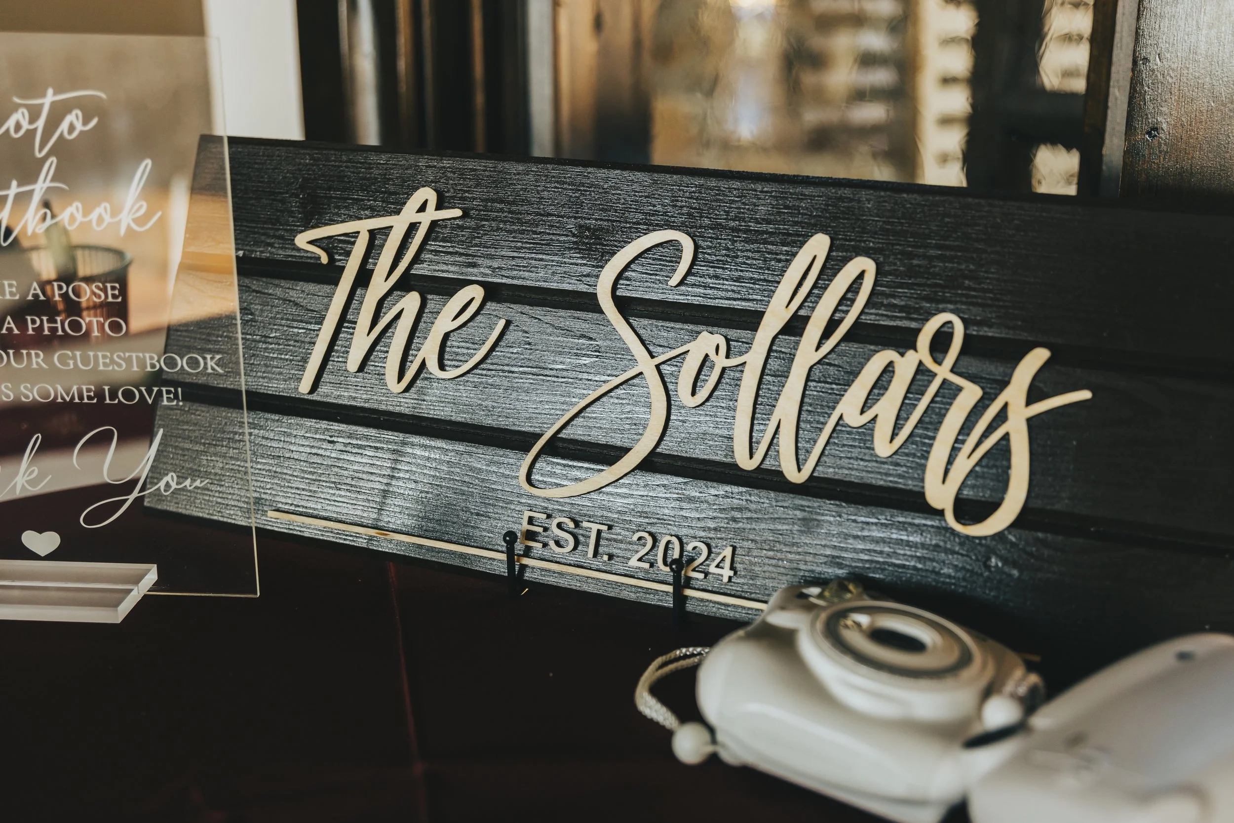 Close-up of a decorative sign that reads "the Salas" with "EST. 2024" beneath it, leaning against a wooden backdrop. Part of a clear acrylic sign with white cursive text and a small white heart symbol also visible.