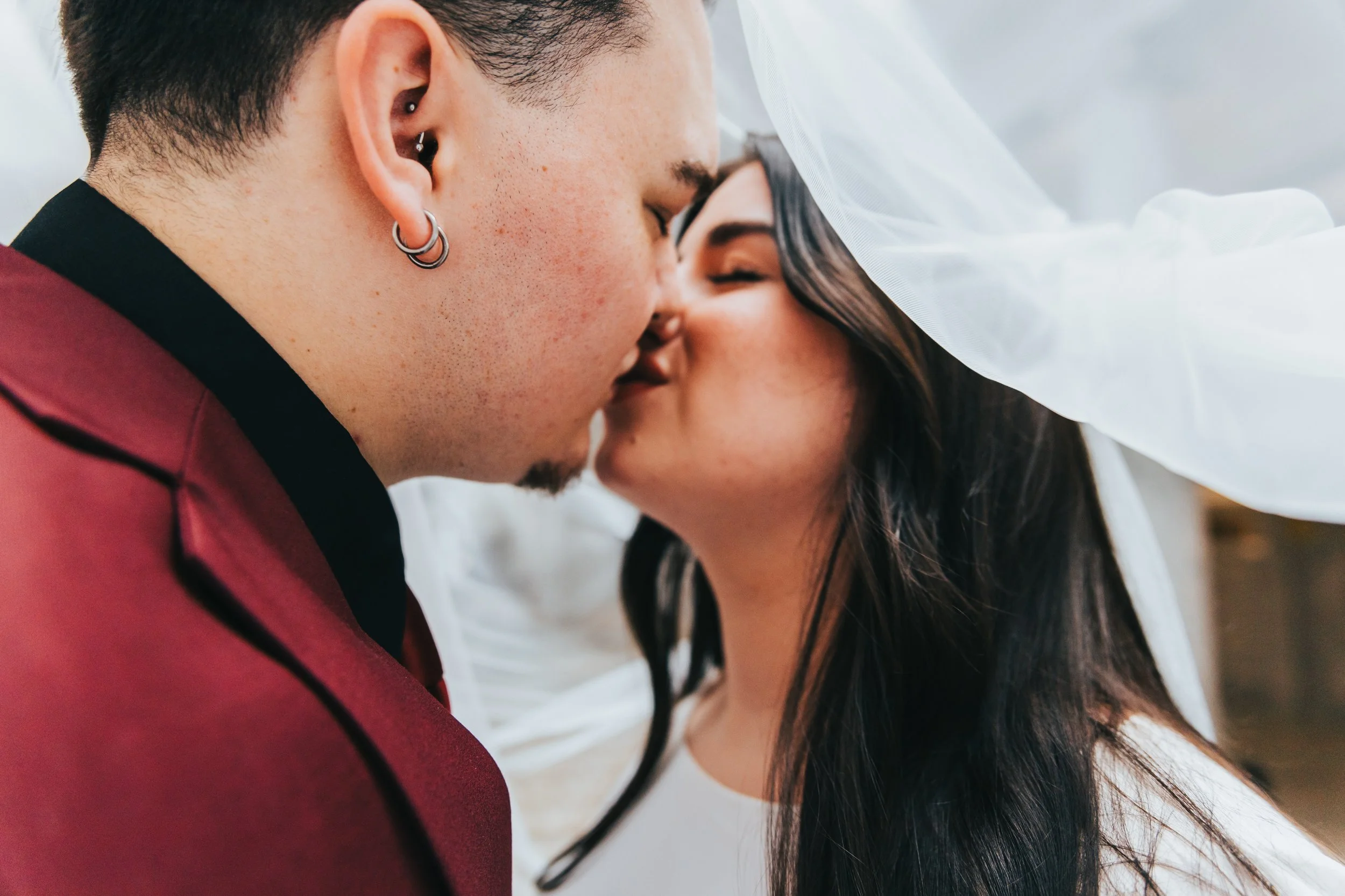A close-up photo of a couple sharing an intimate kiss, their faces pressed closely together, with a white veil visible in the background.