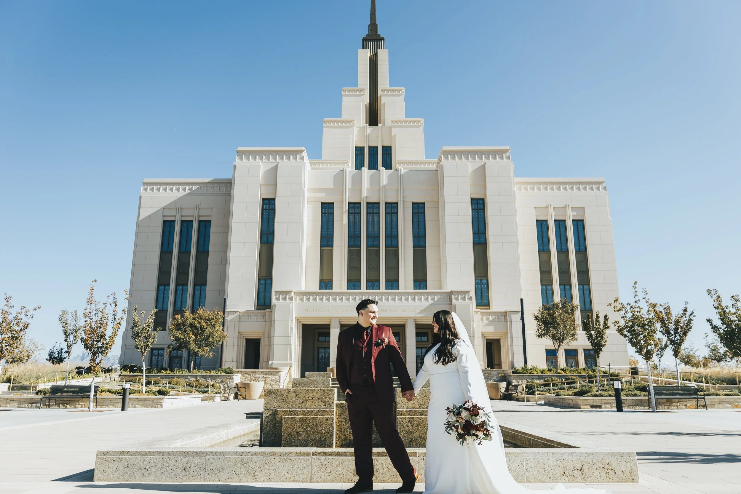 A bride and groom holding hands and smiling outside a large building resembling a temple with a spire, in clear blue sky weather.