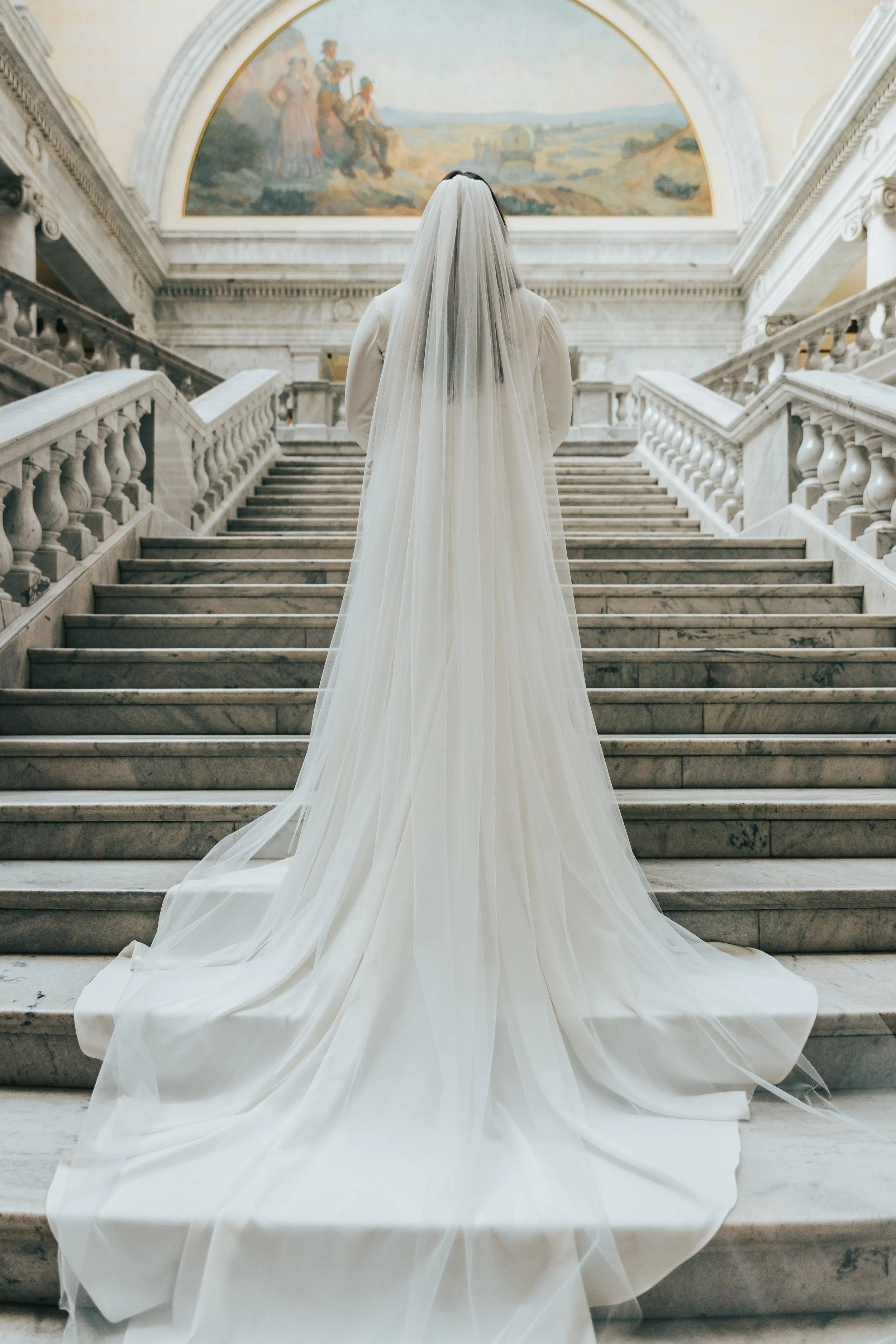 A bride standing on marble stairs inside a building with classical architecture, wearing a long white wedding gown with a veil, facing away from the camera.