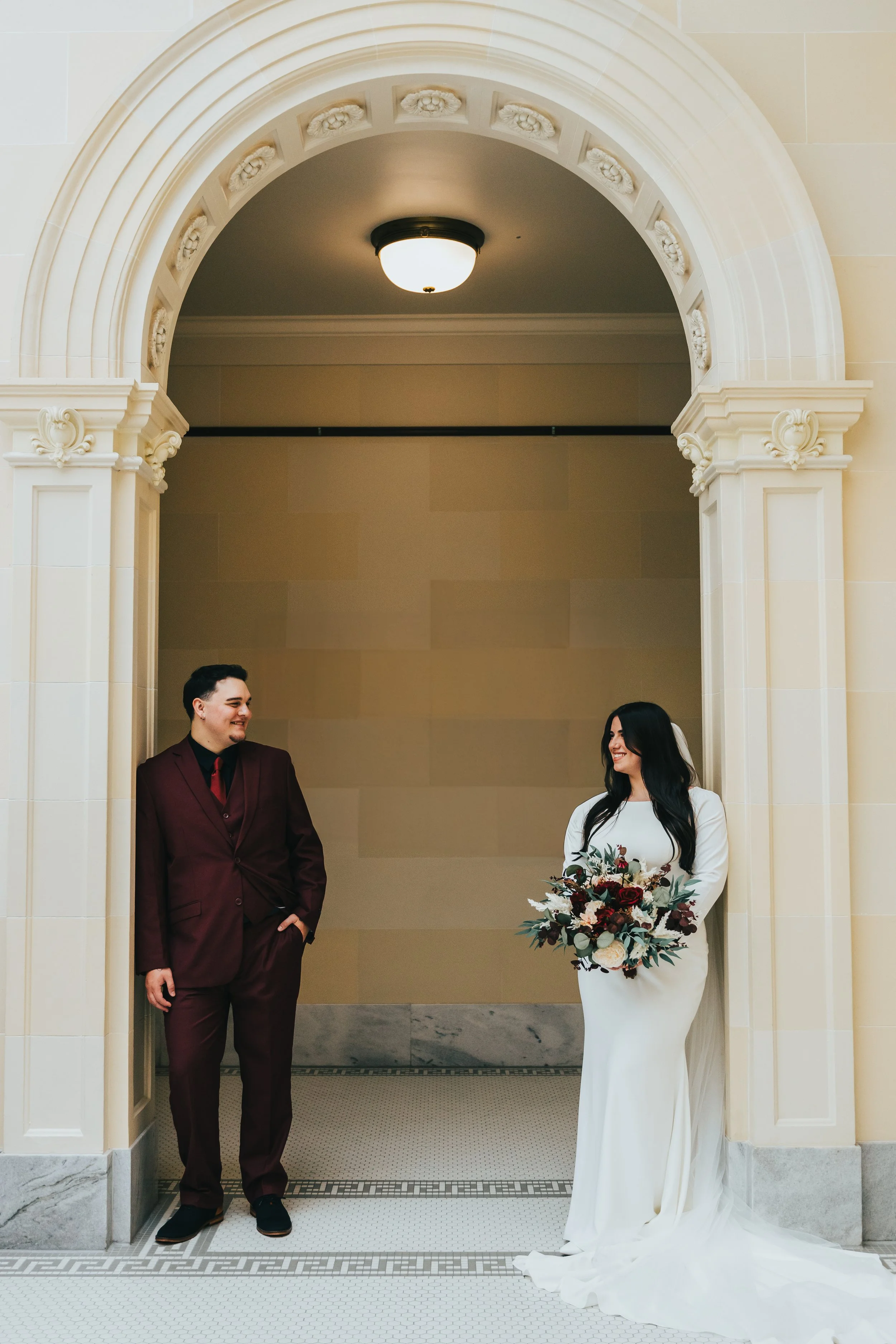 A bride in a white wedding dress holding a bouquet of flowers and a groom in a burgundy suit standing under a decorative archway in an indoor venue.