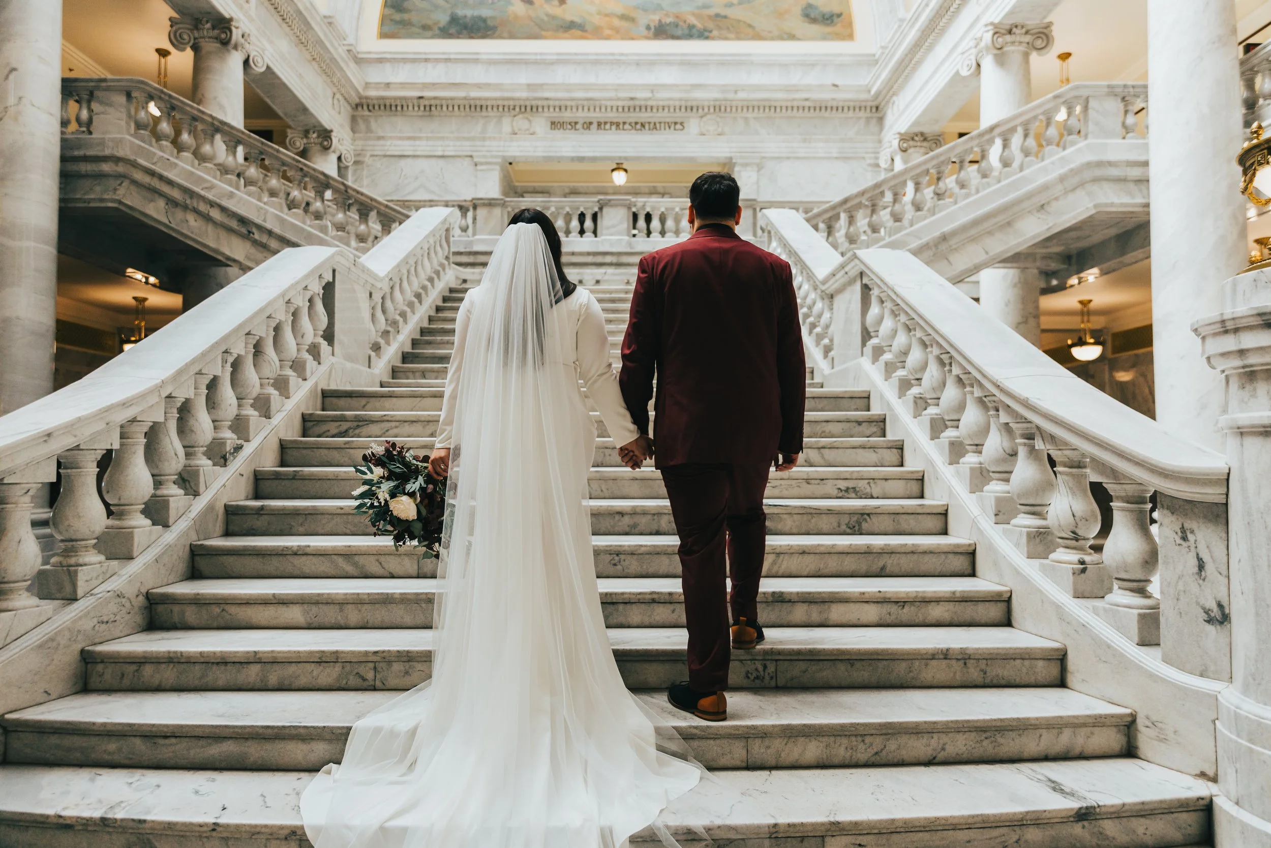 A bride and groom walking up marble stairs in a grand building, holding hands, with the bride wearing a white gown and veil, and the groom in a dark red suit. The building has ornate marble columns and a painting on the ceiling.