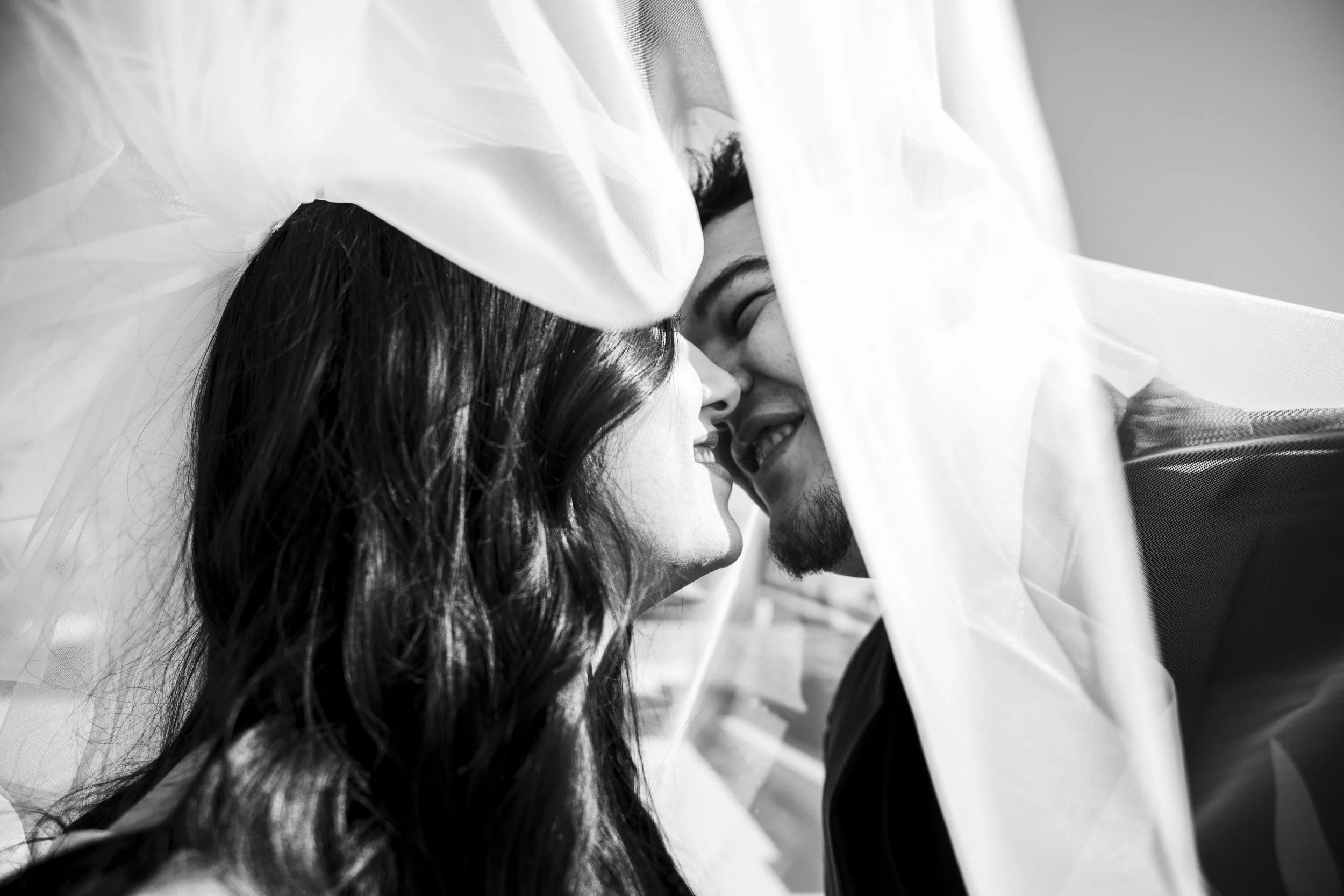 A black and white photo of a couple with their foreheads touching, smiling, partially covered by a translucent veil or fabric.