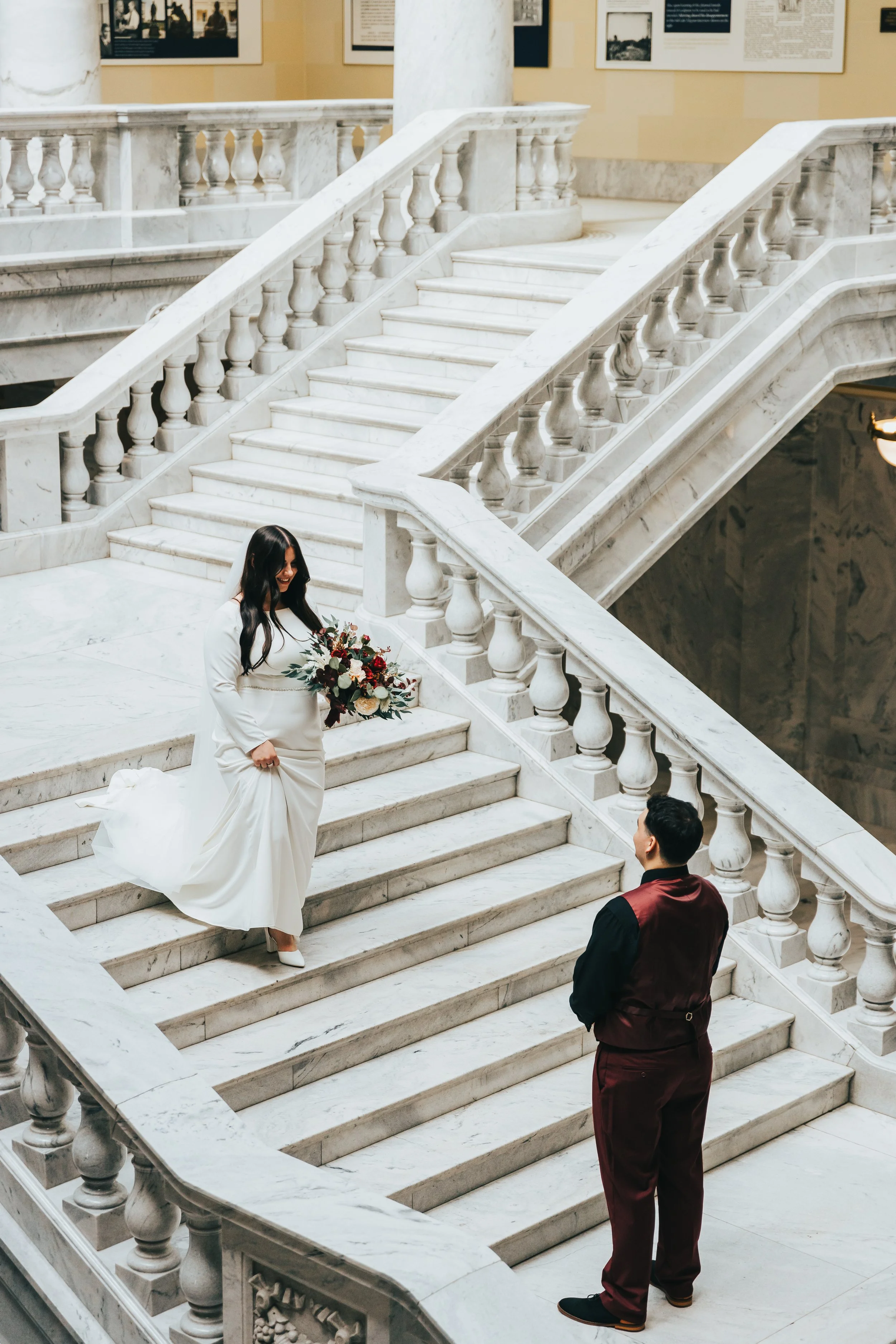 A woman in a white wedding dress holding a bouquet, standing on a marble staircase inside a building, smiling at a man in maroon and black attire, who is looking up at her. The staircase has white marble railings and the background features framed pi