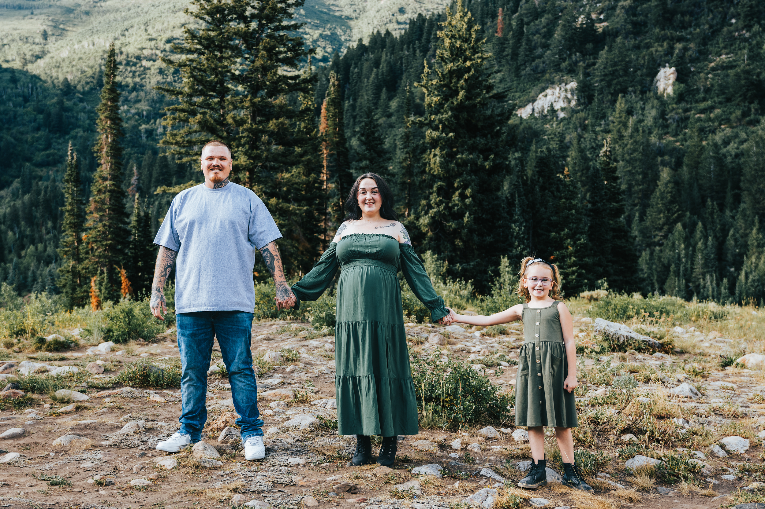 A family of four holding hands in a mountainous forest area, with tall pine trees and rocky terrain.