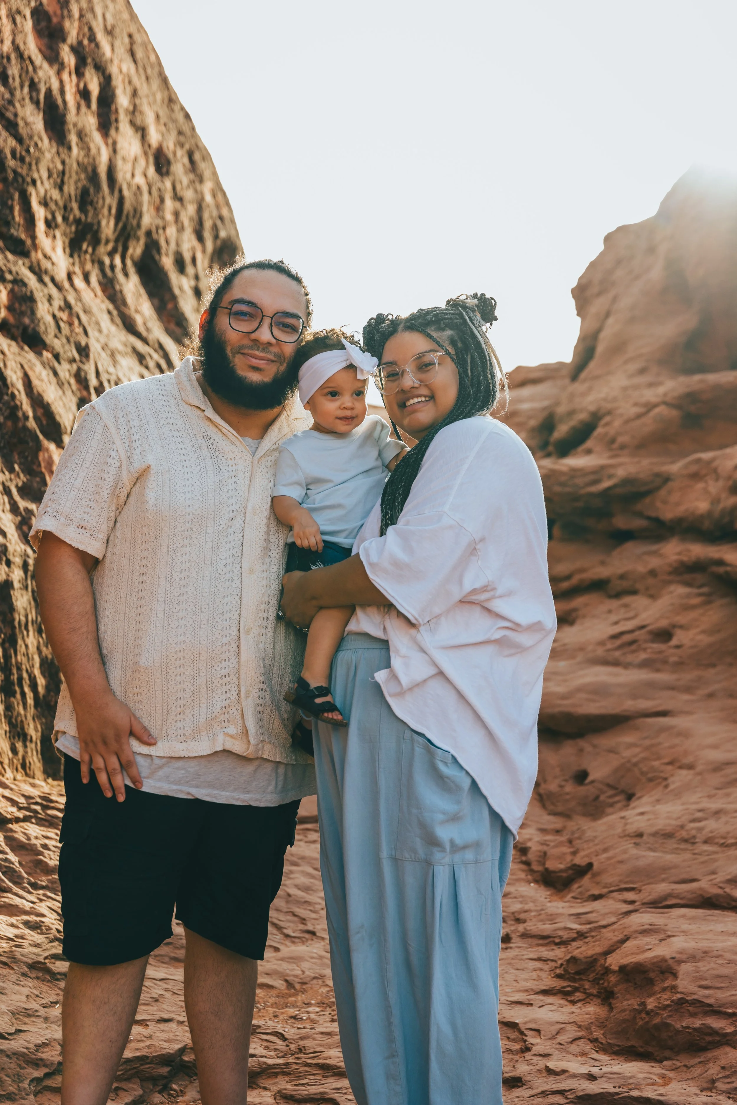 A family of three standing together outdoors in a rocky desert landscape with large rocks and cliffs, smiling for the camera during sunset.