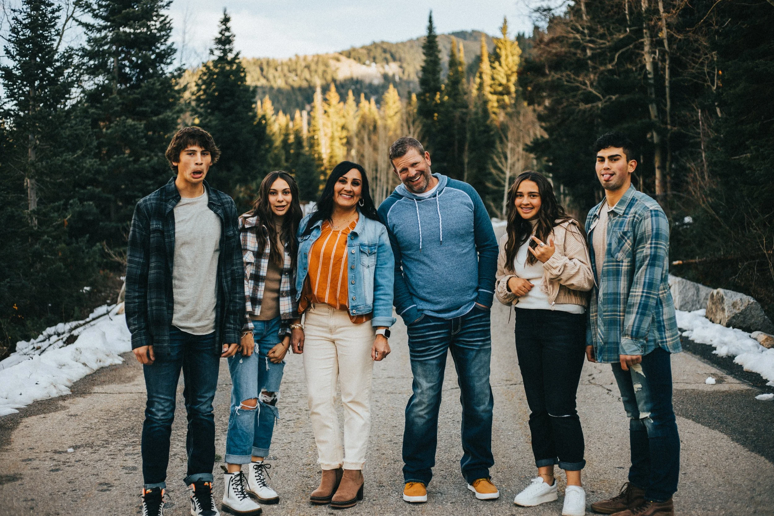 A group of six people, three men and three women, standing outdoors on a mountain trail next to snow, with evergreen trees and mountain scenery in the background. They are smiling and posing for the photo.