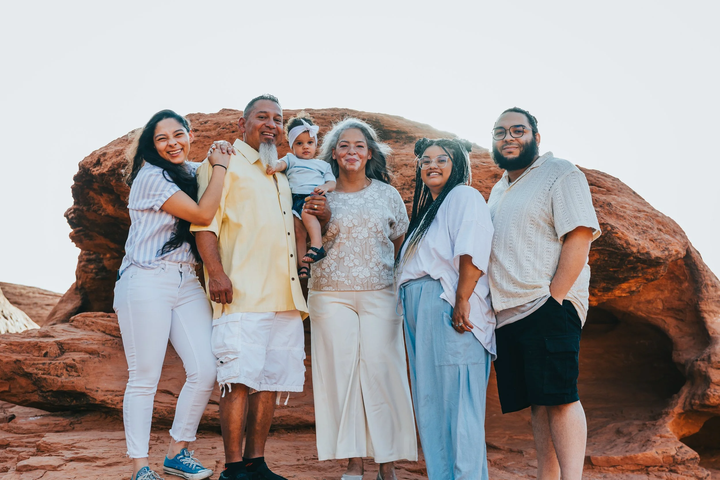 A diverse group of six people, including a child, standing in front of red rock formations, smiling outdoors during daytime.
