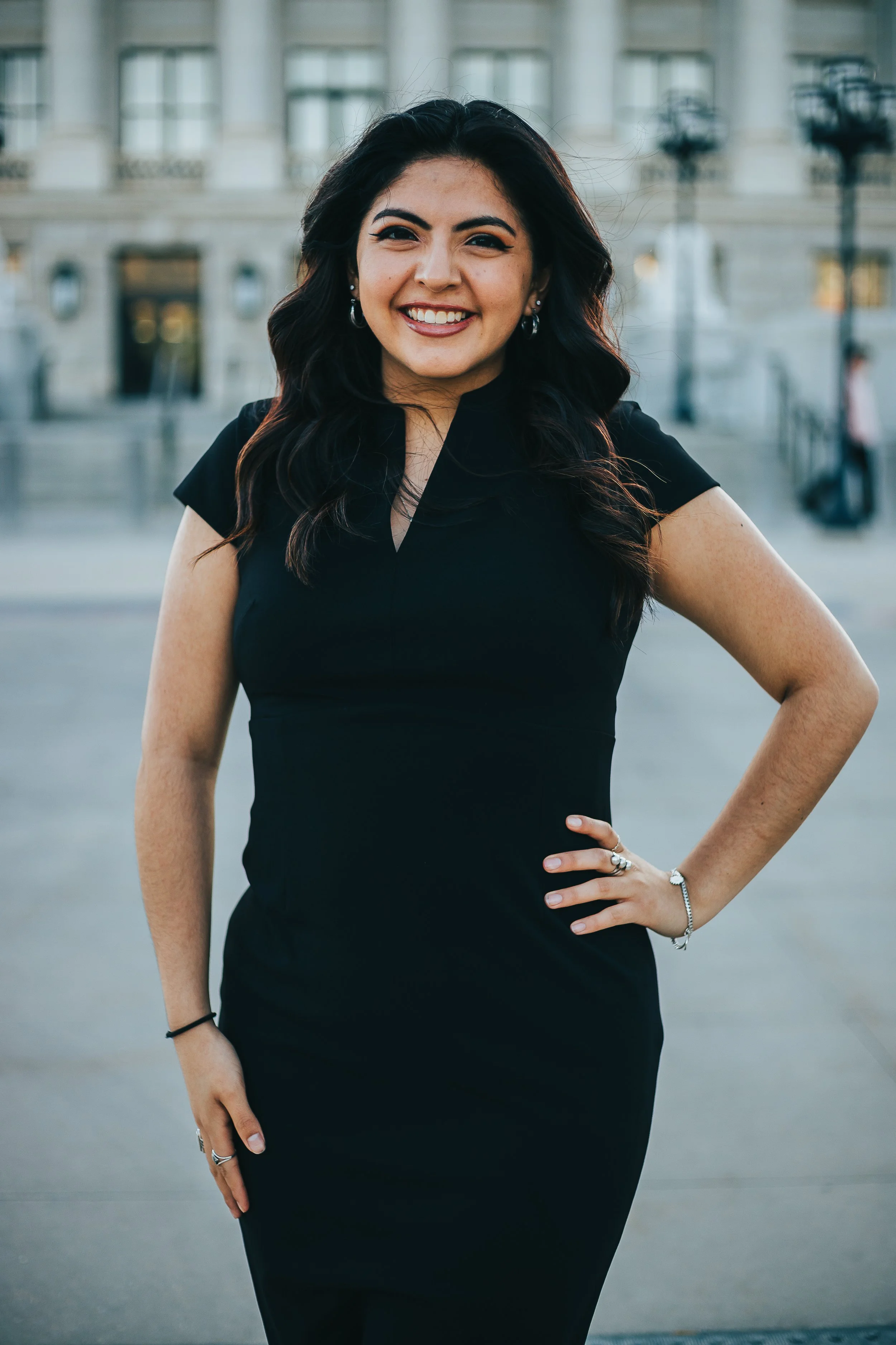 A woman with dark hair, smiling, wearing a black dress and jewelry, standing outdoors in front of a building with stairs and lamps.