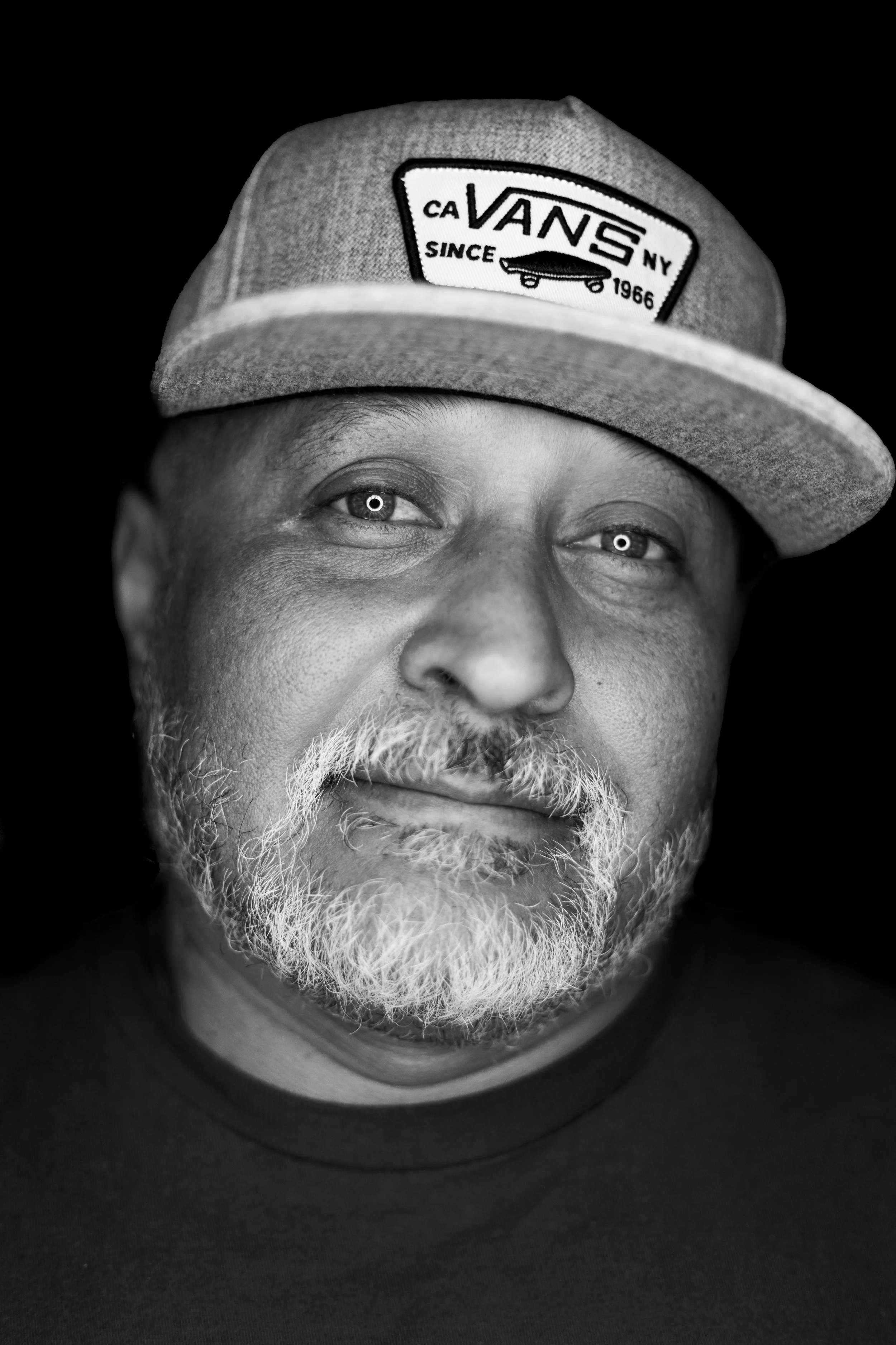 A black-and-white close-up photo of a man wearing a Vans trucker hat with a patch on the front and looking directly at the camera.