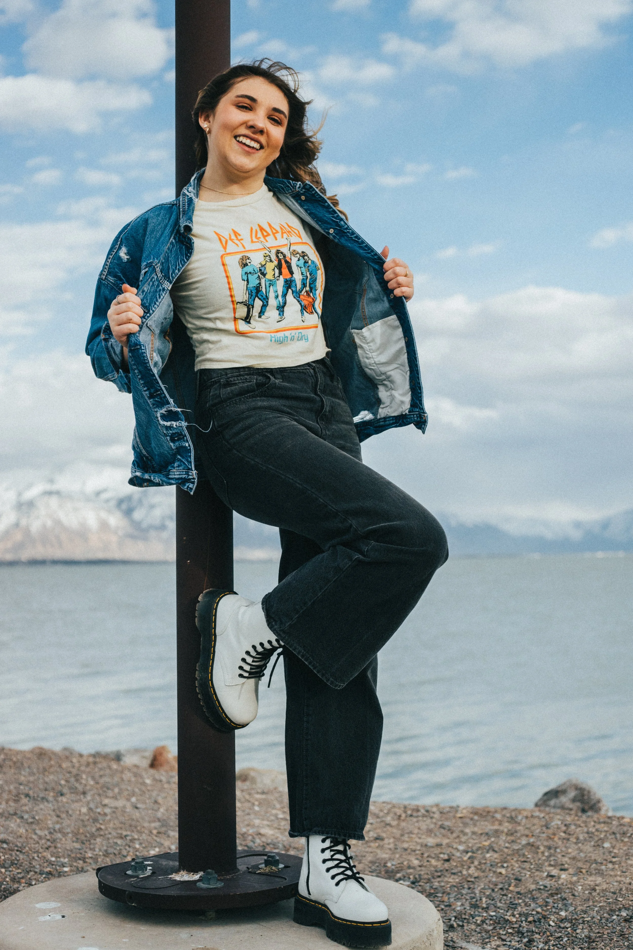 A young woman smiling, standing outdoors by a body of water with mountains in the background. She wears a Def Leppard T-shirt, a denim jacket, black jeans, and white combat boots, holding the jacket open.