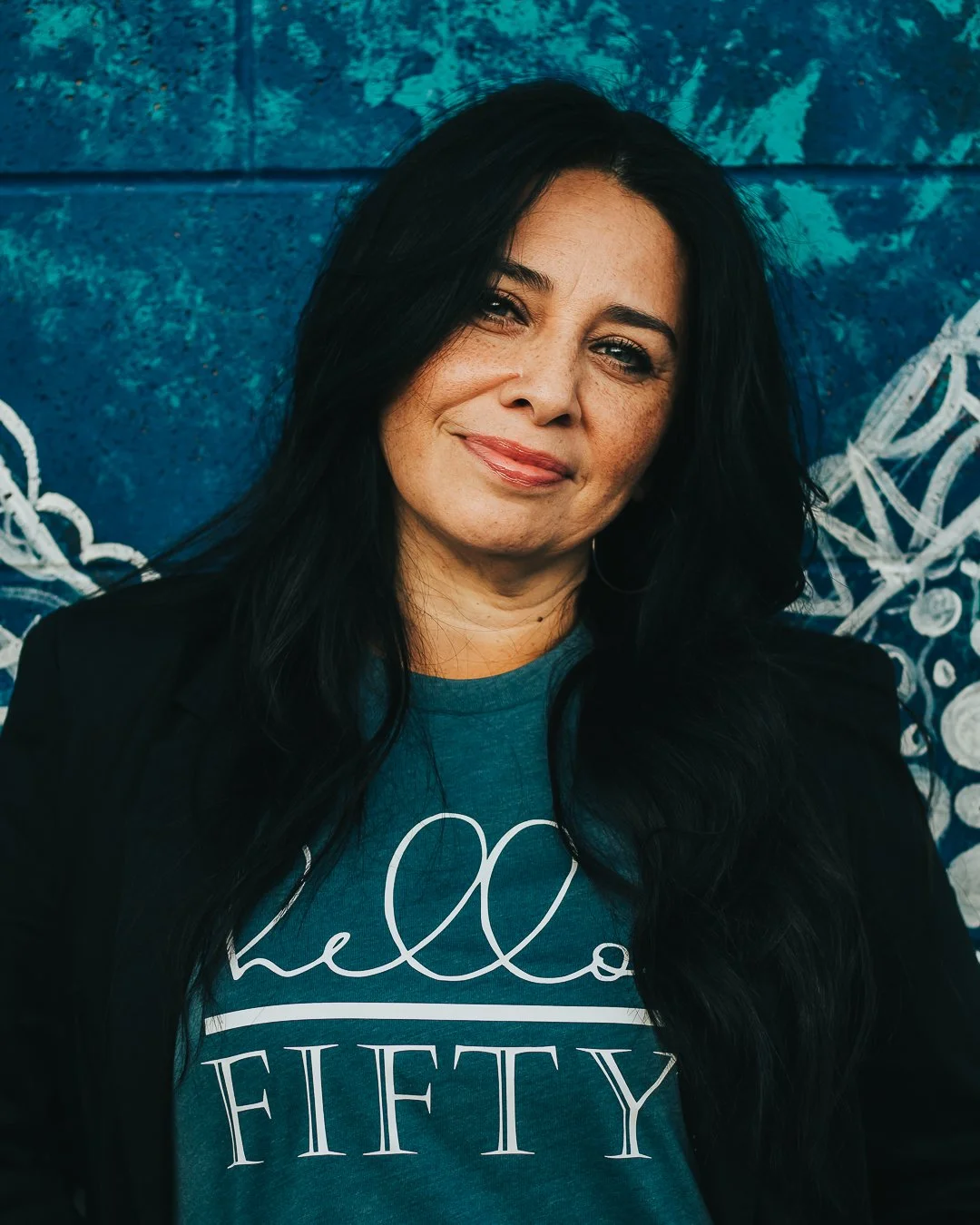 A woman with long black hair wearing a blue shirt and black jacket, standing in front of a blue mural with white abstract designs.