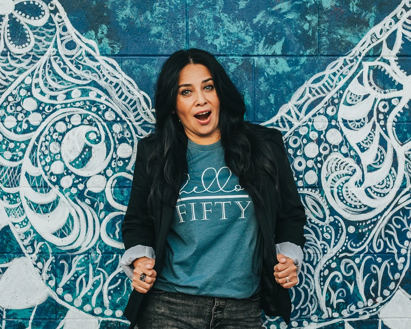 A woman with long dark hair standing in front of a vibrant blue mural with white decorative patterns. She is wearing a blue shirt that says 'Fifty', a black jacket, and has an expressive look with her mouth open and fists slightly raised.