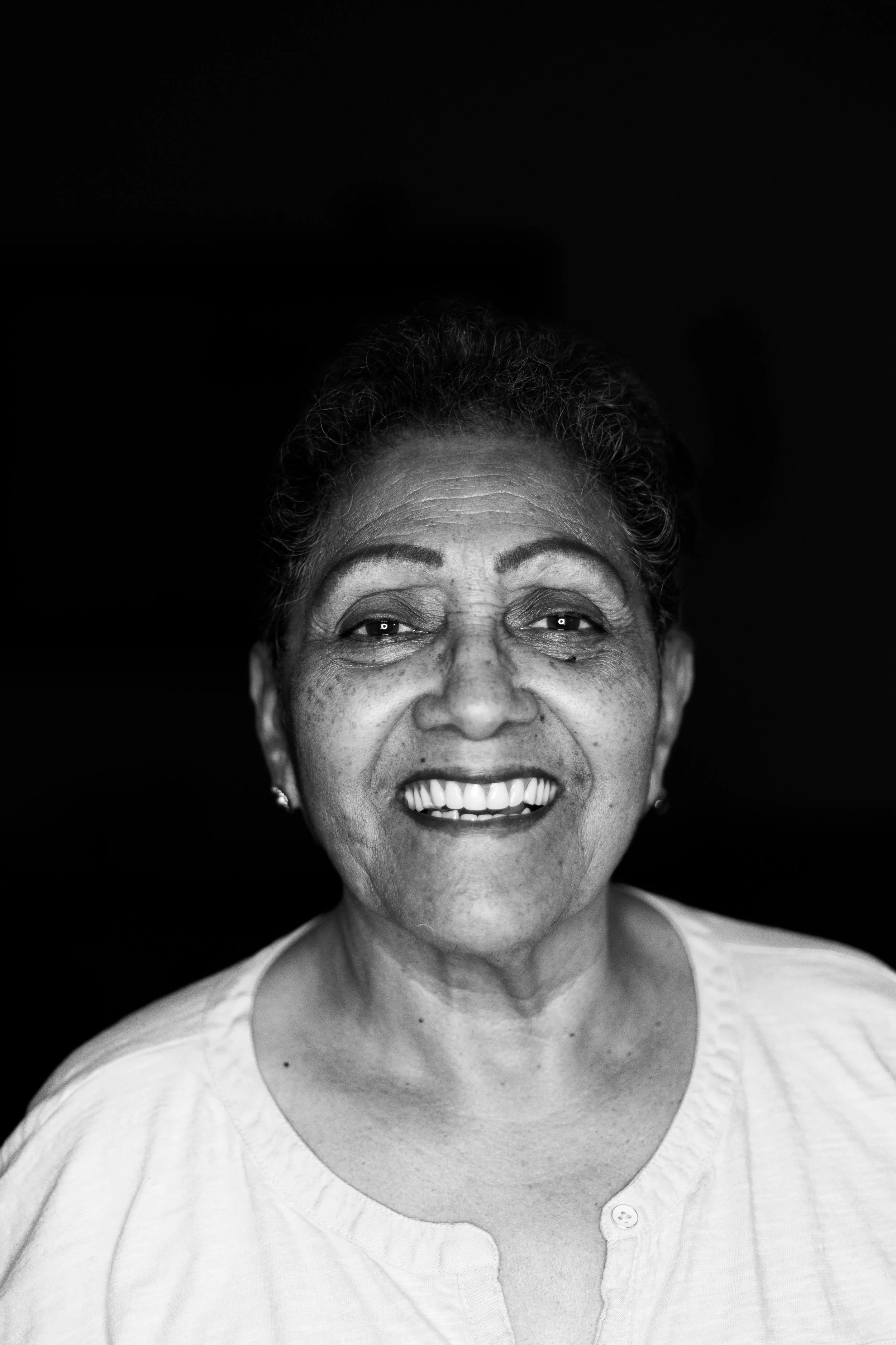 Close-up black and white photograph of an smiling older woman with curly hair, wearing a buttoned shirt, against a dark background.