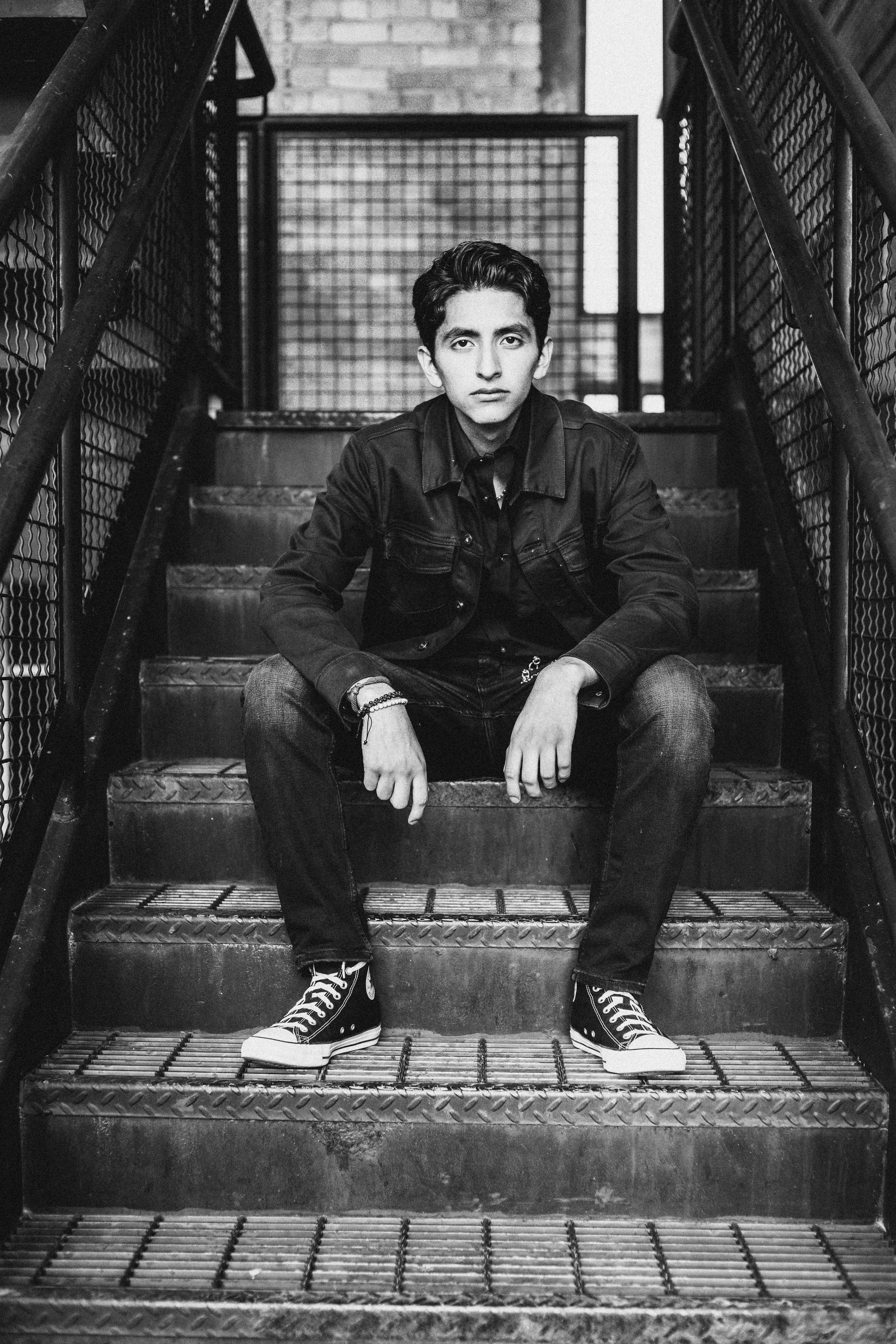 A black and white photo of a young man sitting on metal stairs in an urban setting, wearing a denim jacket, jeans, and sneakers, with a metal grid and brick wall in the background.