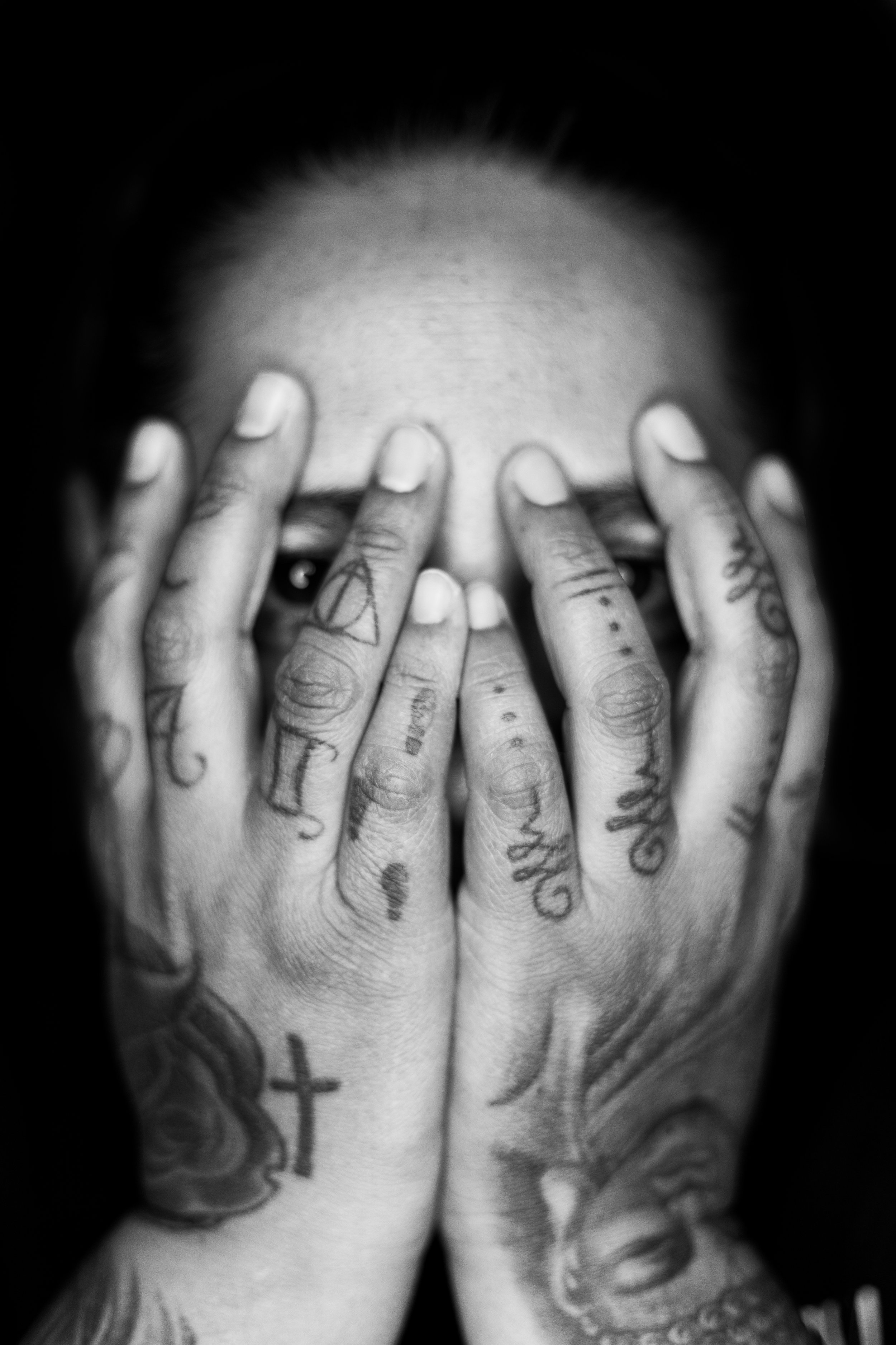 Black and white photo of hands covering a person's face, with tattoos on fingers and hands.