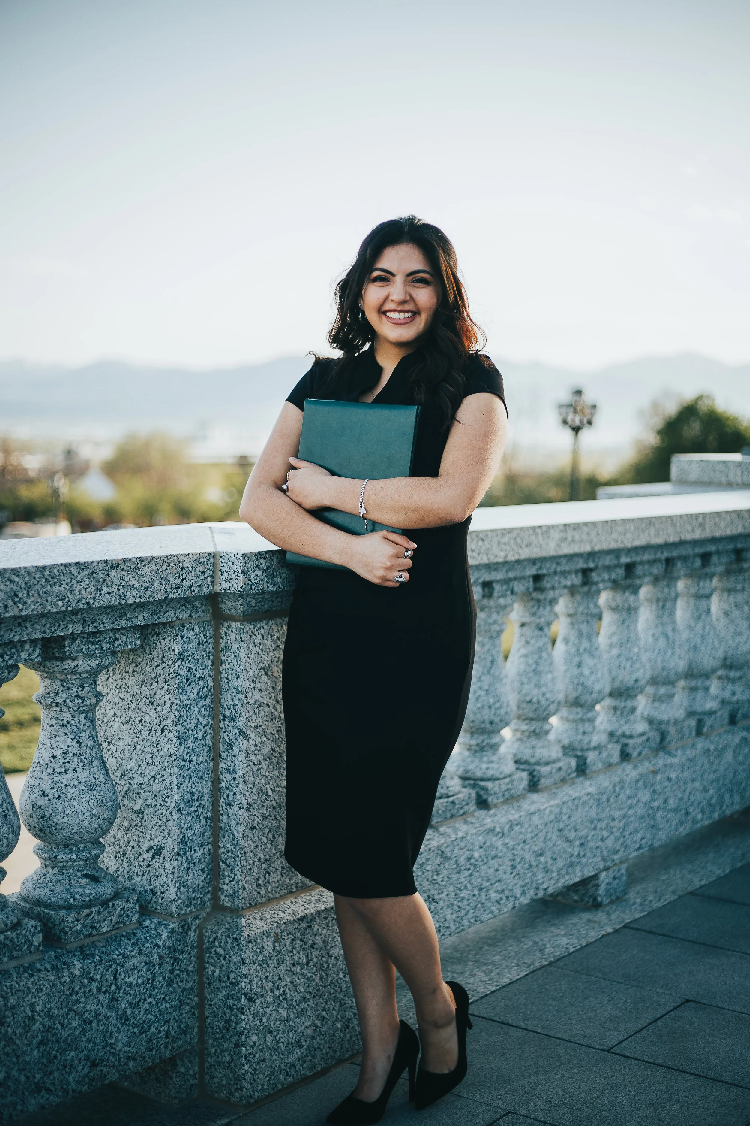 A woman in a black dress with black high heels standing outdoors near a stone railing, holding a teal folder, smiling at the camera, with hills and trees in the background during late afternoon or early evening.