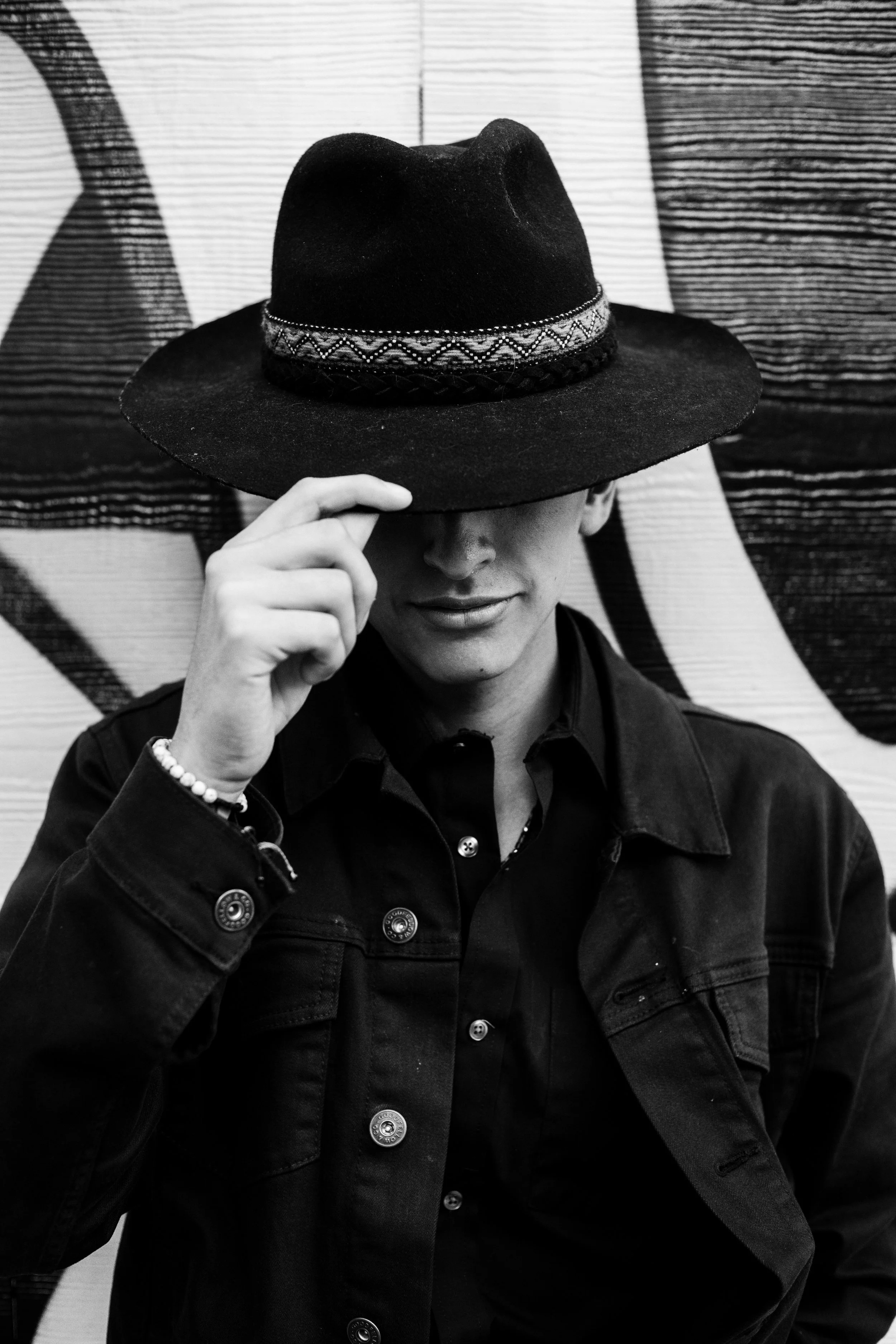 A person wearing a dark jacket and partly hidden by a wide-brimmed hat, standing against a tapestry with horizontal and vertical striped patterns, holding the brim of the hat with one hand.