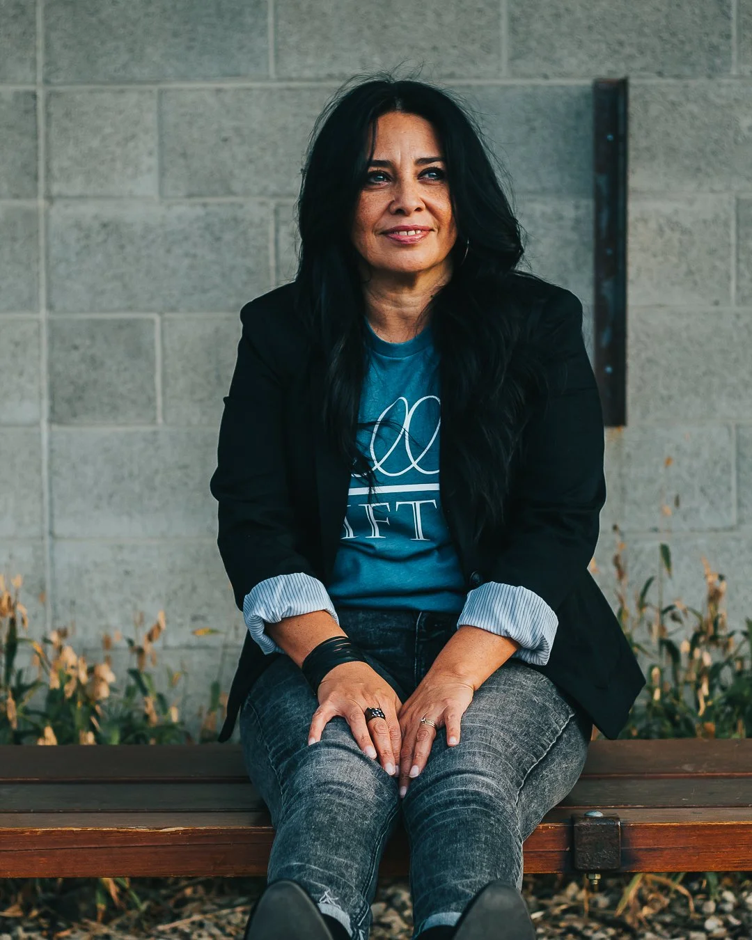 A woman with long black hair sitting on a wooden bench against a gray brick wall, wearing a black blazer, a blue graphic t-shirt, and gray jeans, smiling softly.