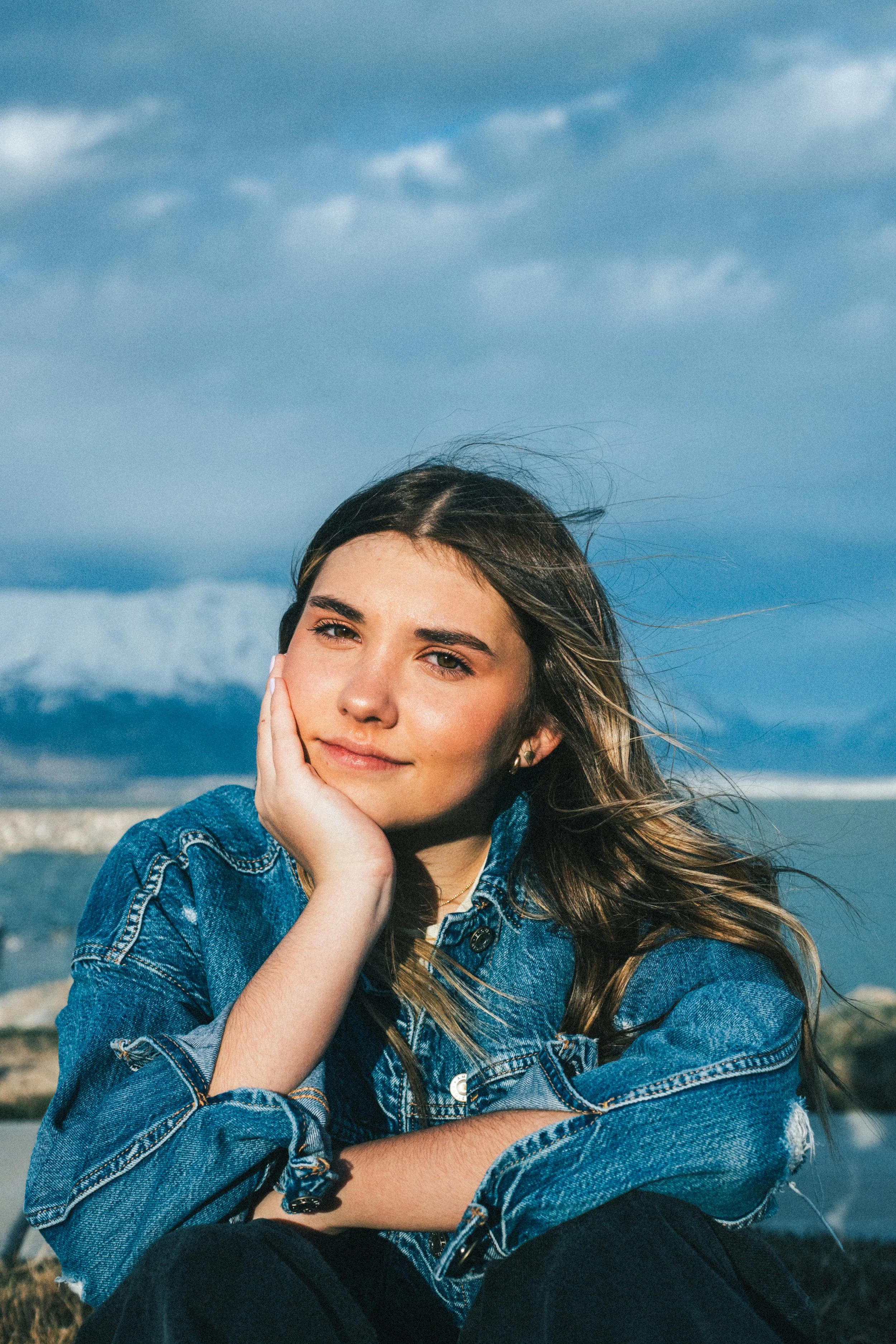 A young woman sitting outdoors near a body of water with a cloudy sky in the background. She has long, wavy hair, is wearing a denim jacket, and has a relaxed, contemplative expression, resting her face on her hand.