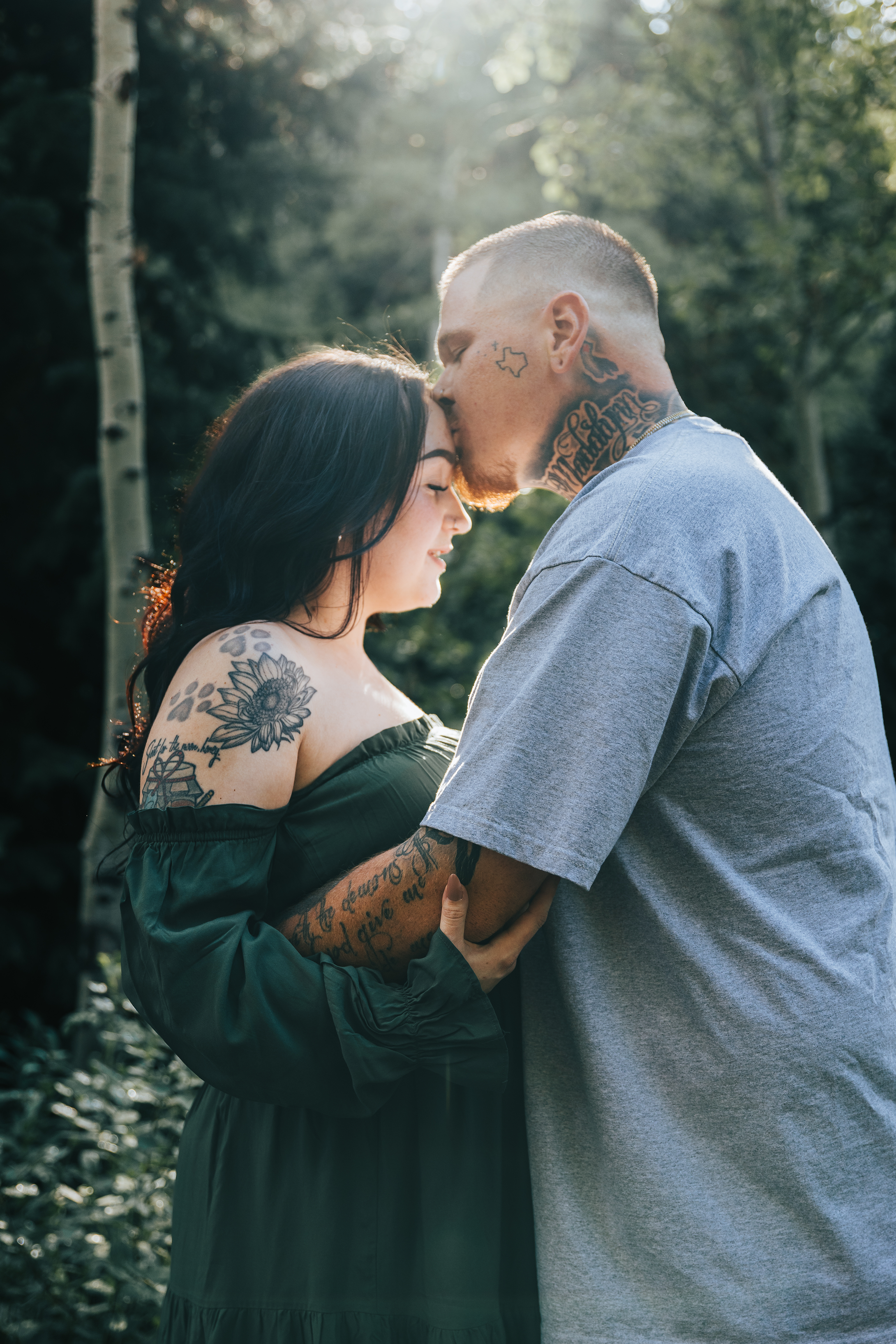 A couple stands outdoors in a wooded area, sharing an intimate moment with foreheads touching, eyes closed. The woman has dark hair, tattoos on her shoulder and arm, and is wearing an off-the-shoulder green dress. The man has short blonde hair and ta