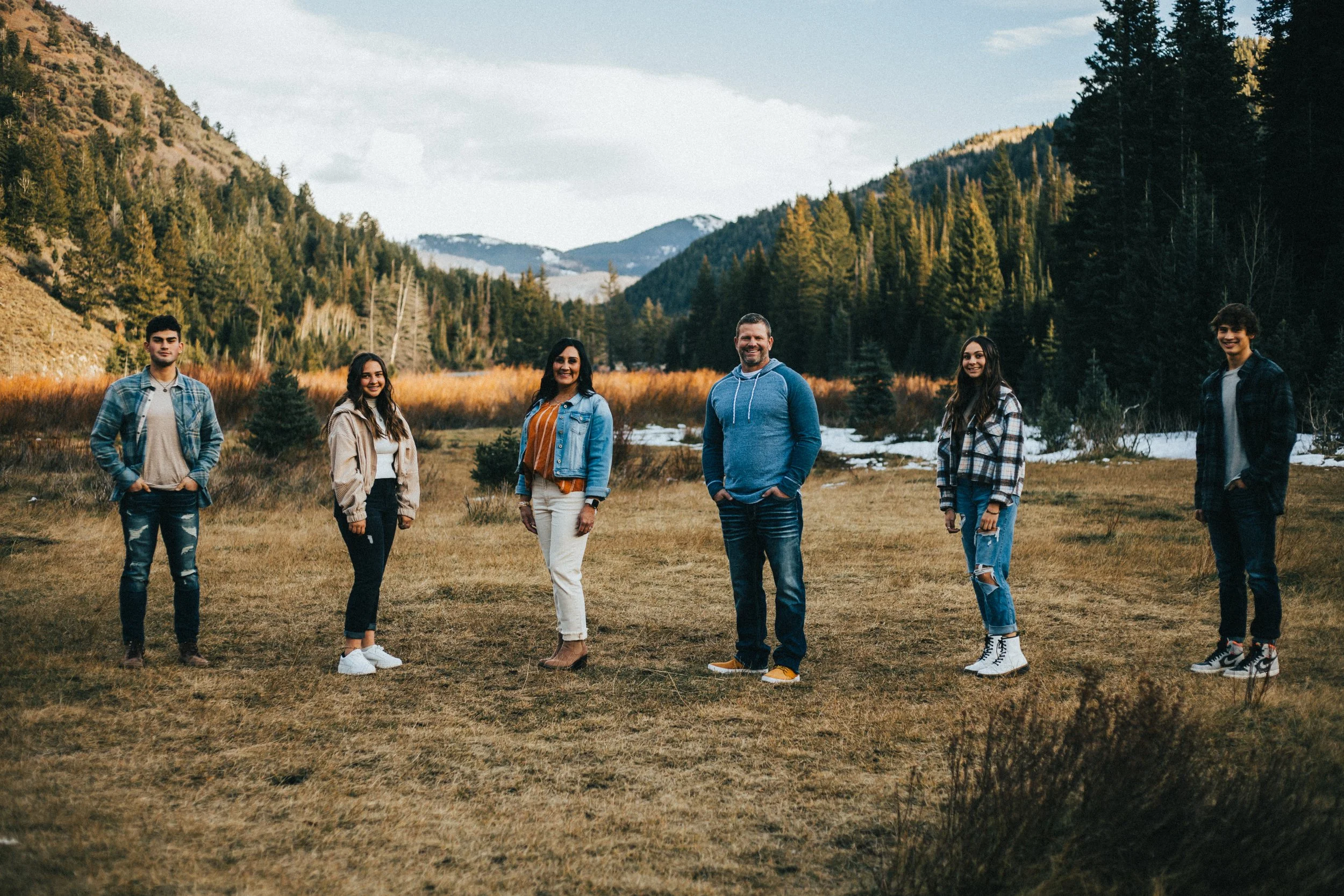 Group of six people standing outdoors in a mountain valley with forested hills and snow-capped peaks in the background.