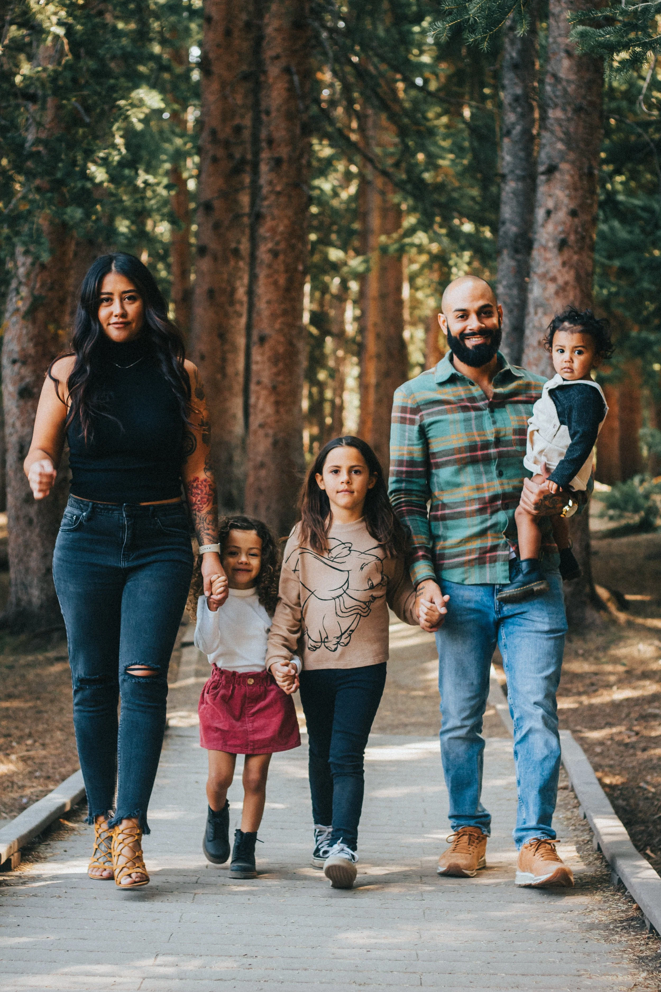A diverse family walking together on a paved trail through a forest with tall trees and green foliage.
